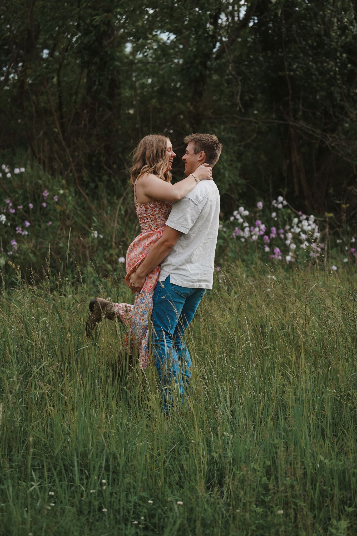 Romantic couple embraces in tall grass meadow with wildflowers and wooded backdrop during sunset.
