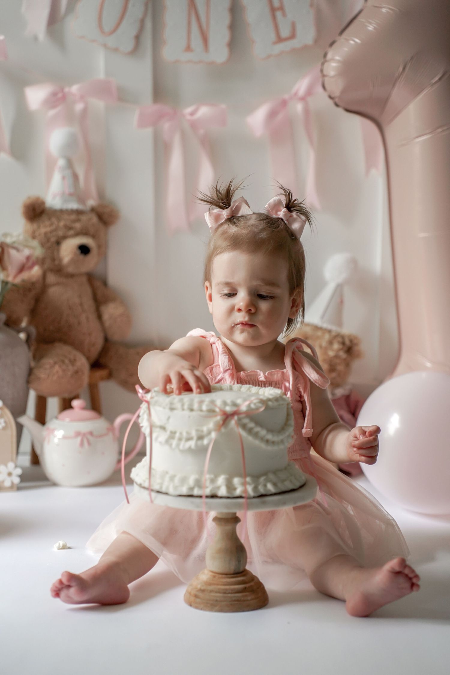 Pink and white first birthday party setup with a frosted cake on a pedestal stand and teddy bear decorations.