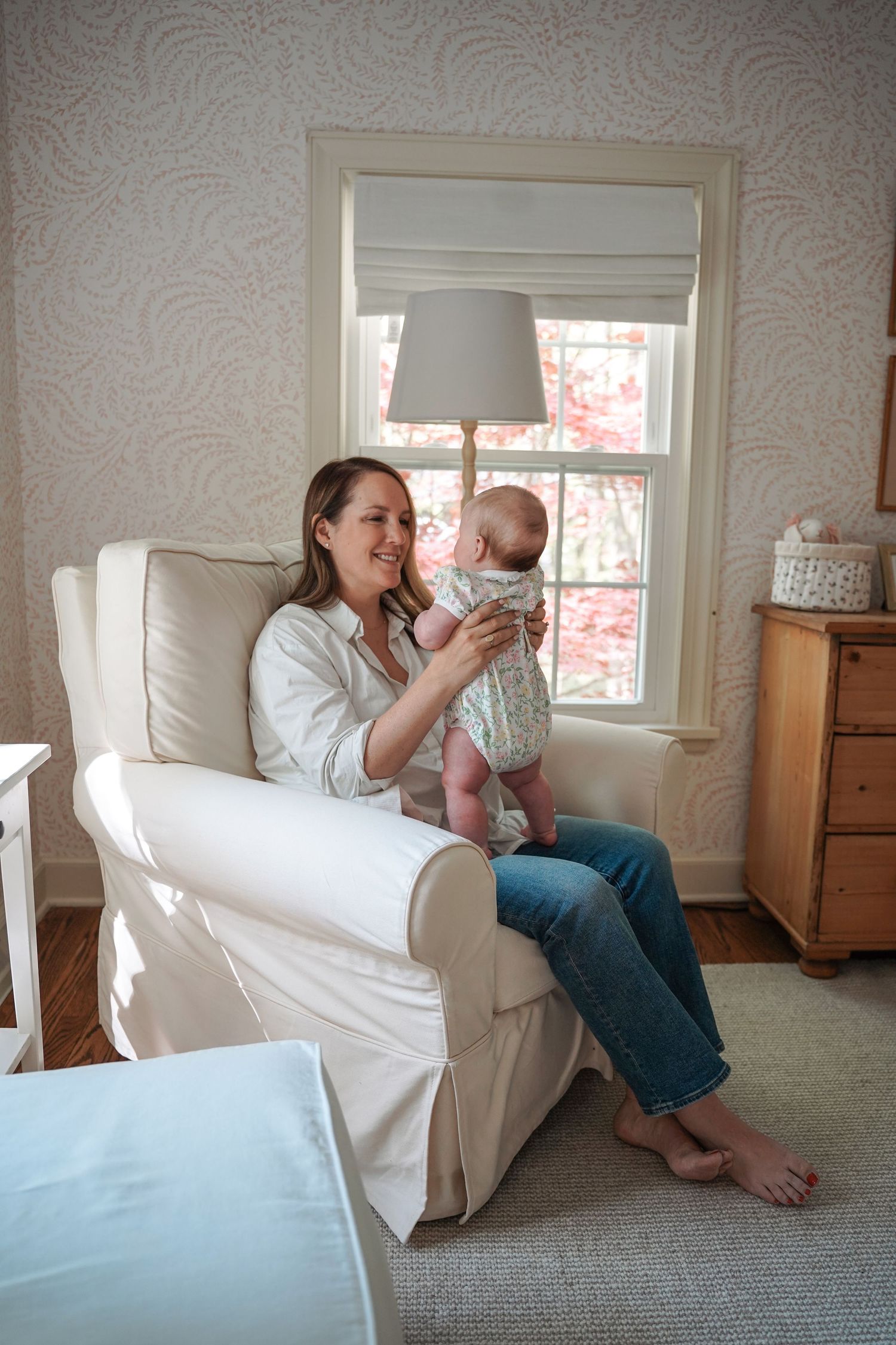 A peaceful moment in a sunlit nursery room with a white armchair, window, lamp and wooden dresser.