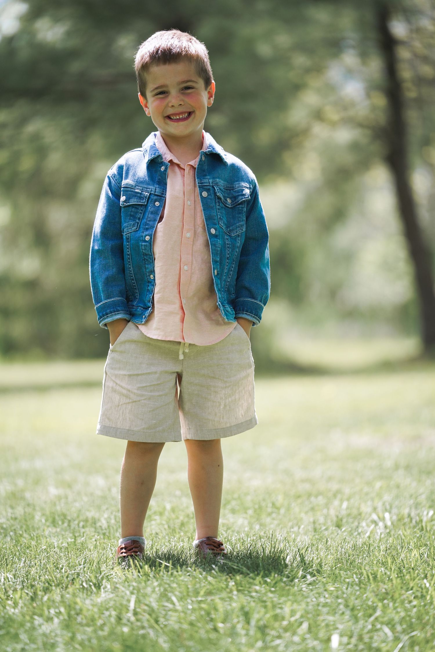 A young child stands on grass wearing a denim jacket, pink shirt and khaki shorts on a sunny day outdoors.