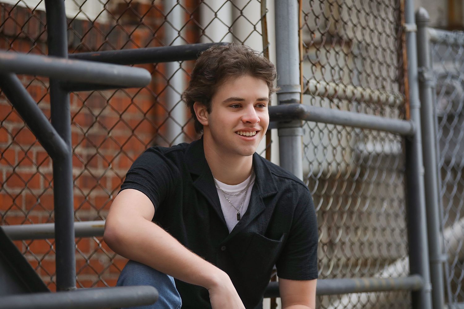 A person in a black button-up shirt sits casually on metal bleachers with a chain link fence and brick wall behind them.
