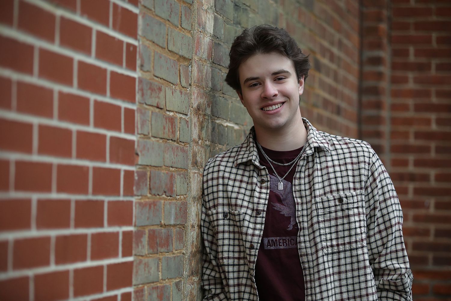 A person in a checkered shirt and burgundy t-shirt stands against a brick wall smiling at the camera.