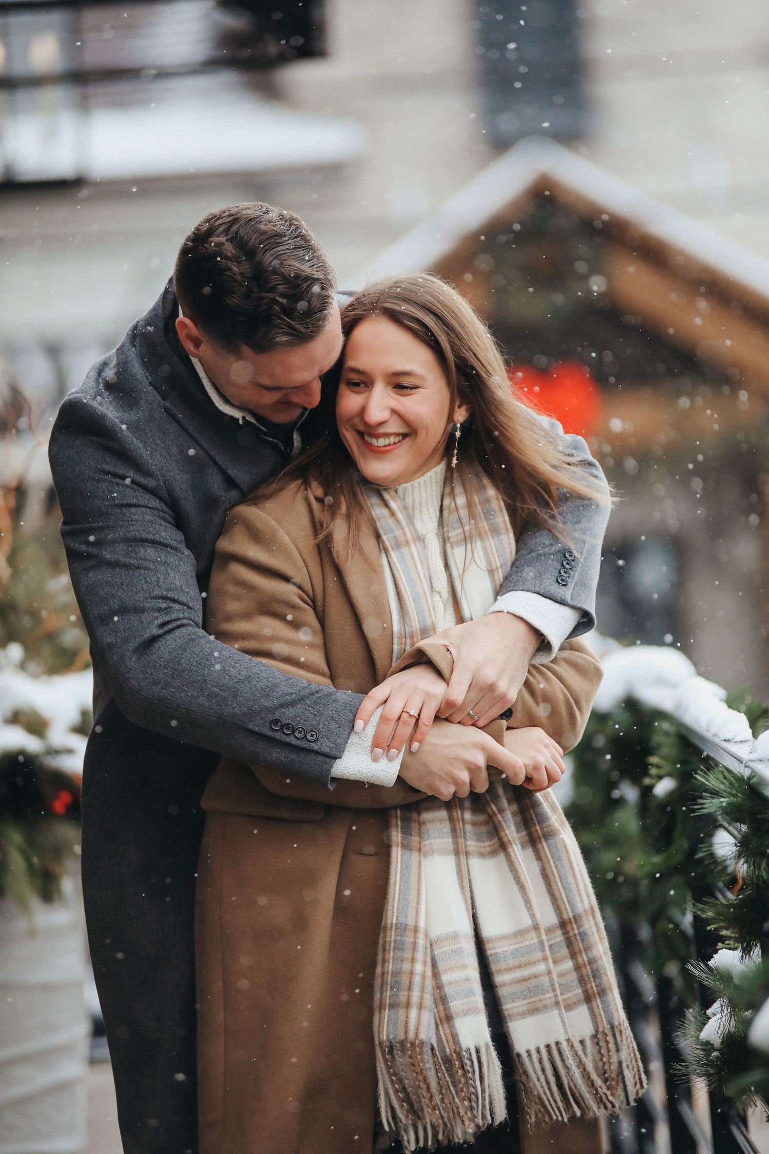A couple shares an intimate moment in the snow while wearing winter coats and a plaid scarf.