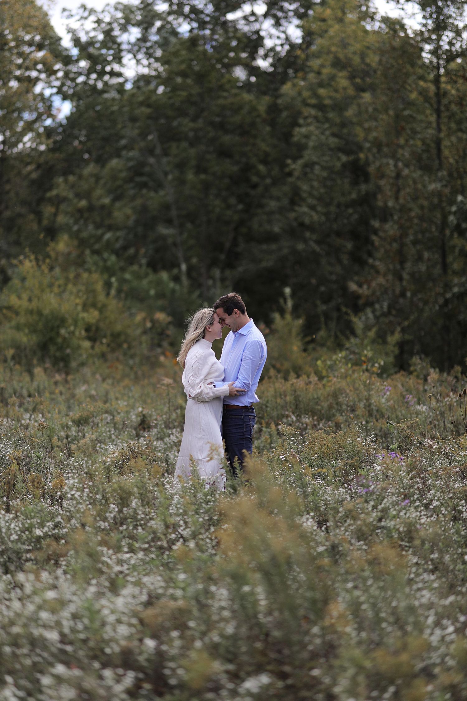 A couple shares an intimate moment in a wildflower meadow surrounded by trees on a peaceful summer evening.