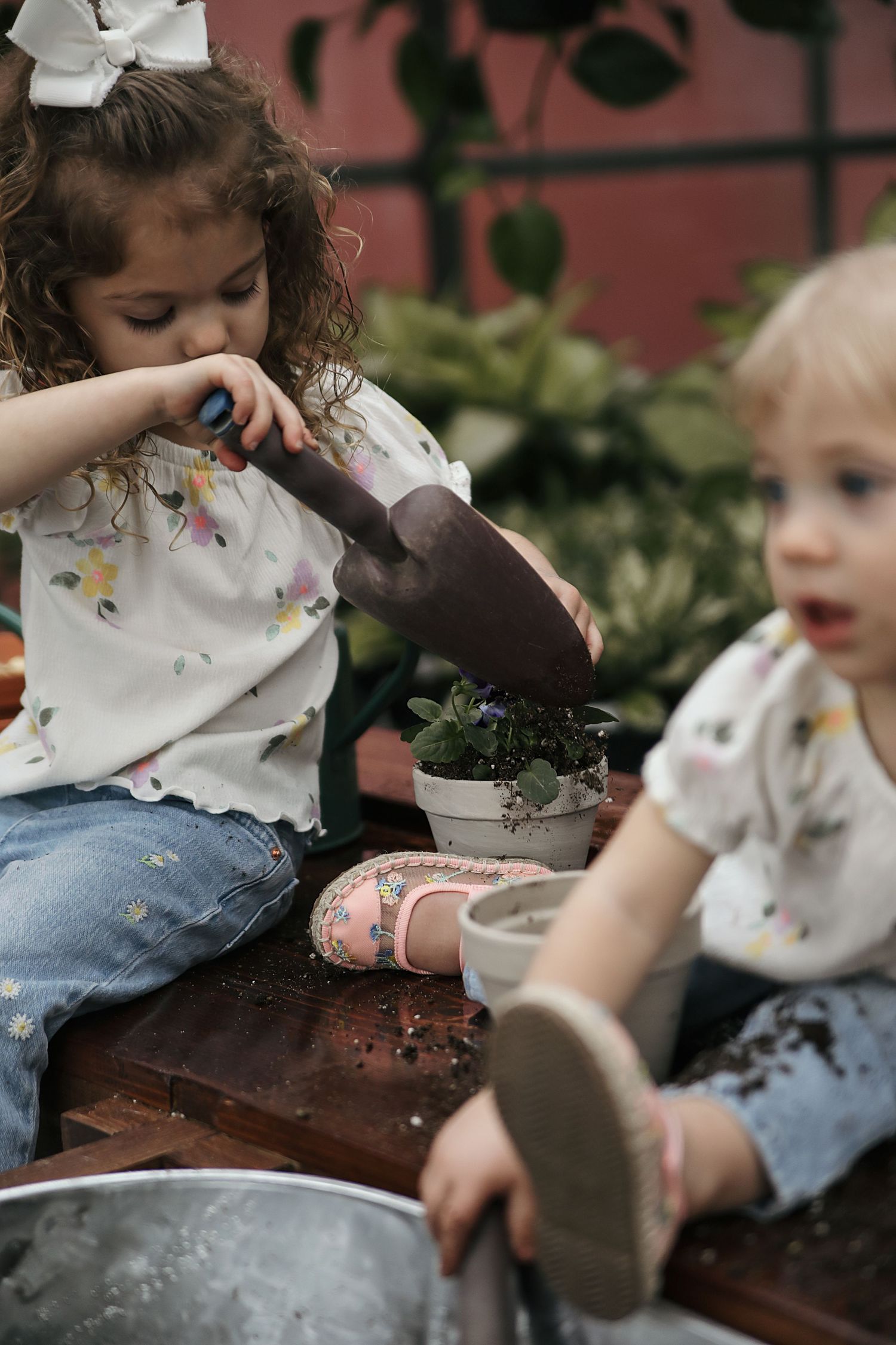 Two children in floral shirts and jeans playing with gardening pots and soil on a wooden surface outdoors.