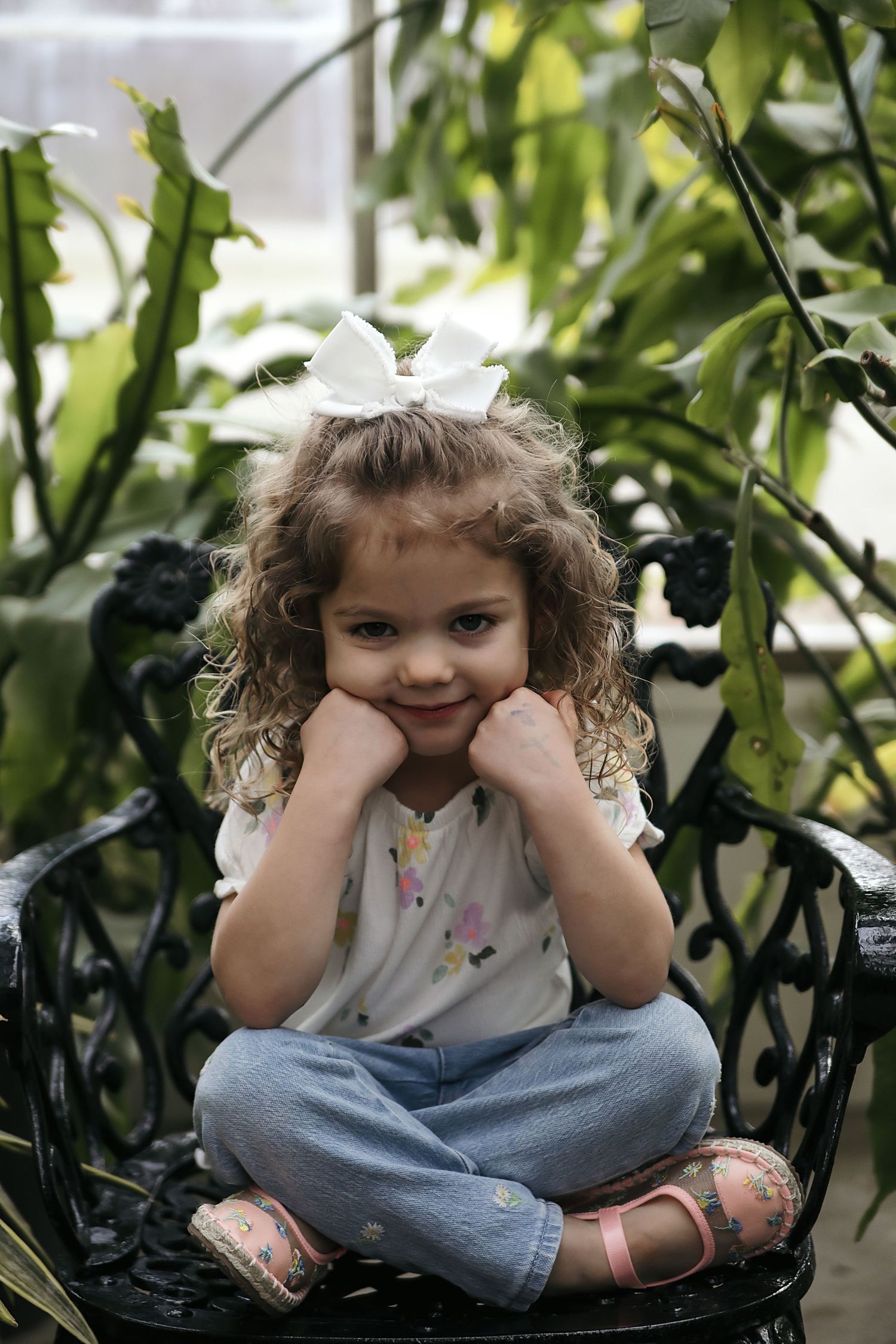 A smiling child in blue jeans sits cross-legged in a lush green garden setting.