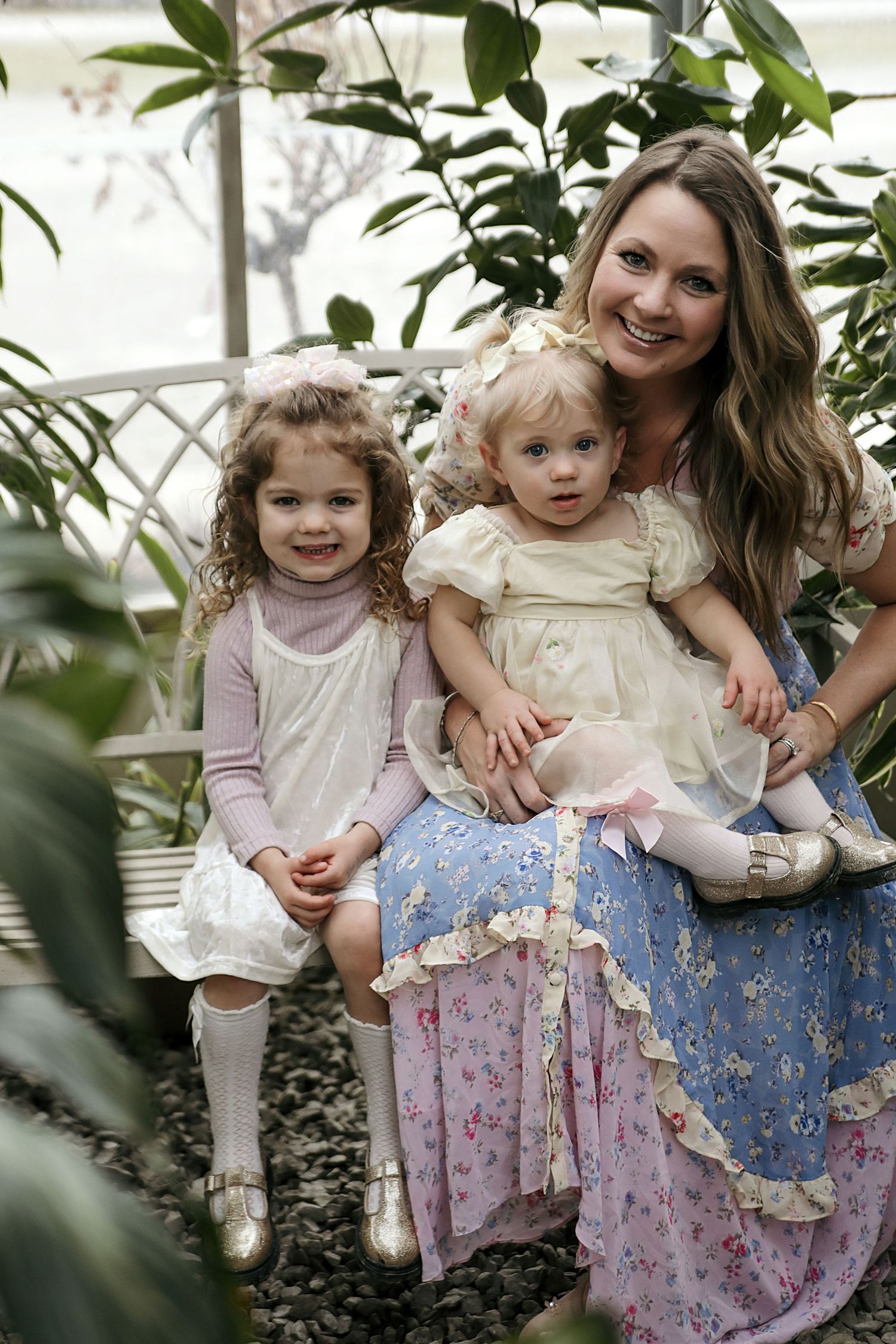 A charming family portrait in a greenhouse setting with lush plants and floral dresses creating a dreamy, natural atmosphere.