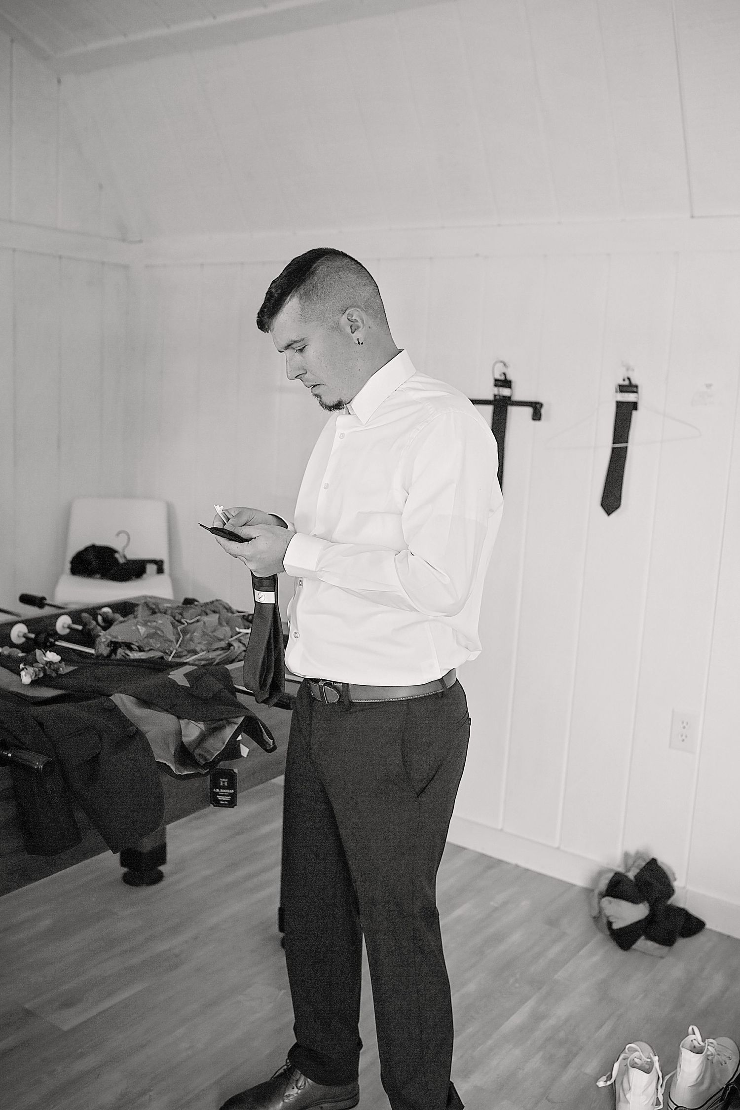 A person in formal attire checks their phone while getting ready in a simple white room.