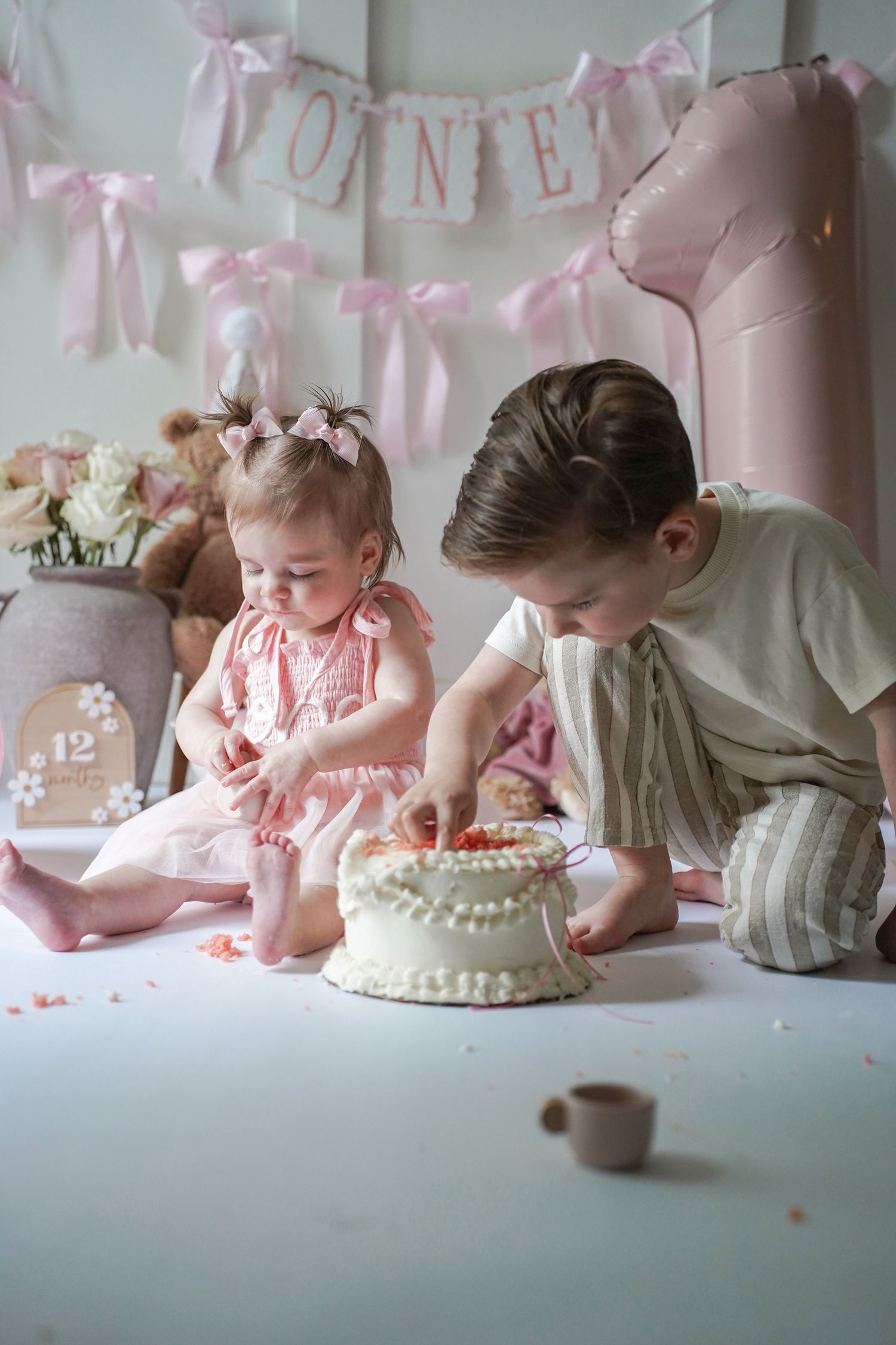 Two children in pink and white celebrate first birthday with cake decorations and pink ribbon garland decorations.