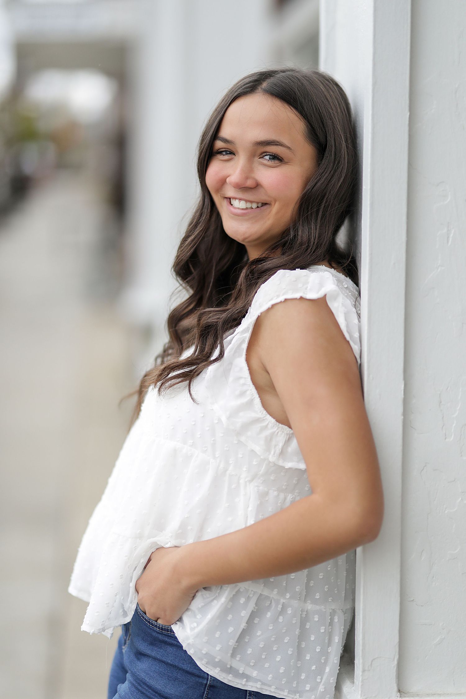 Young woman in white eyelet top and jeans poses playfully against white columns in outdoor portrait series.