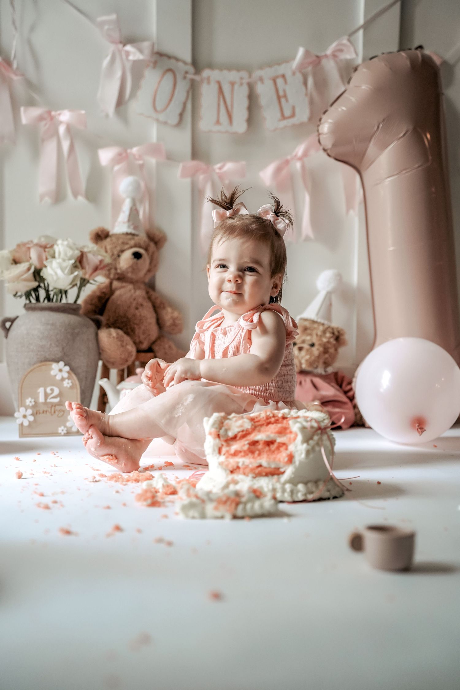 Pink first birthday cake smash photoshoot setup with balloons, banner, teddy bears and frosting-covered cake.
