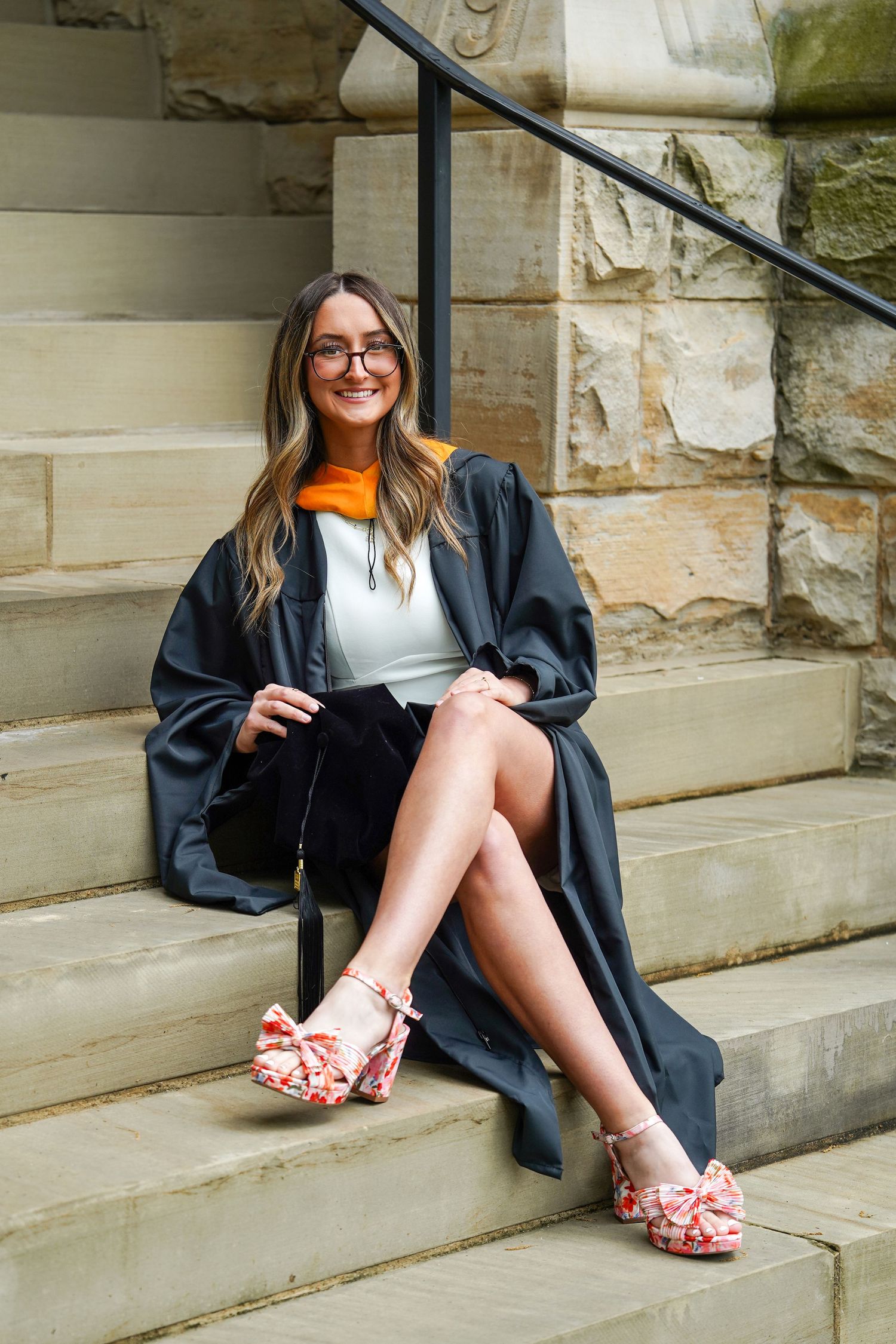 Graduate in black cap and gown sits on stone steps wearing floral shoes and glasses.