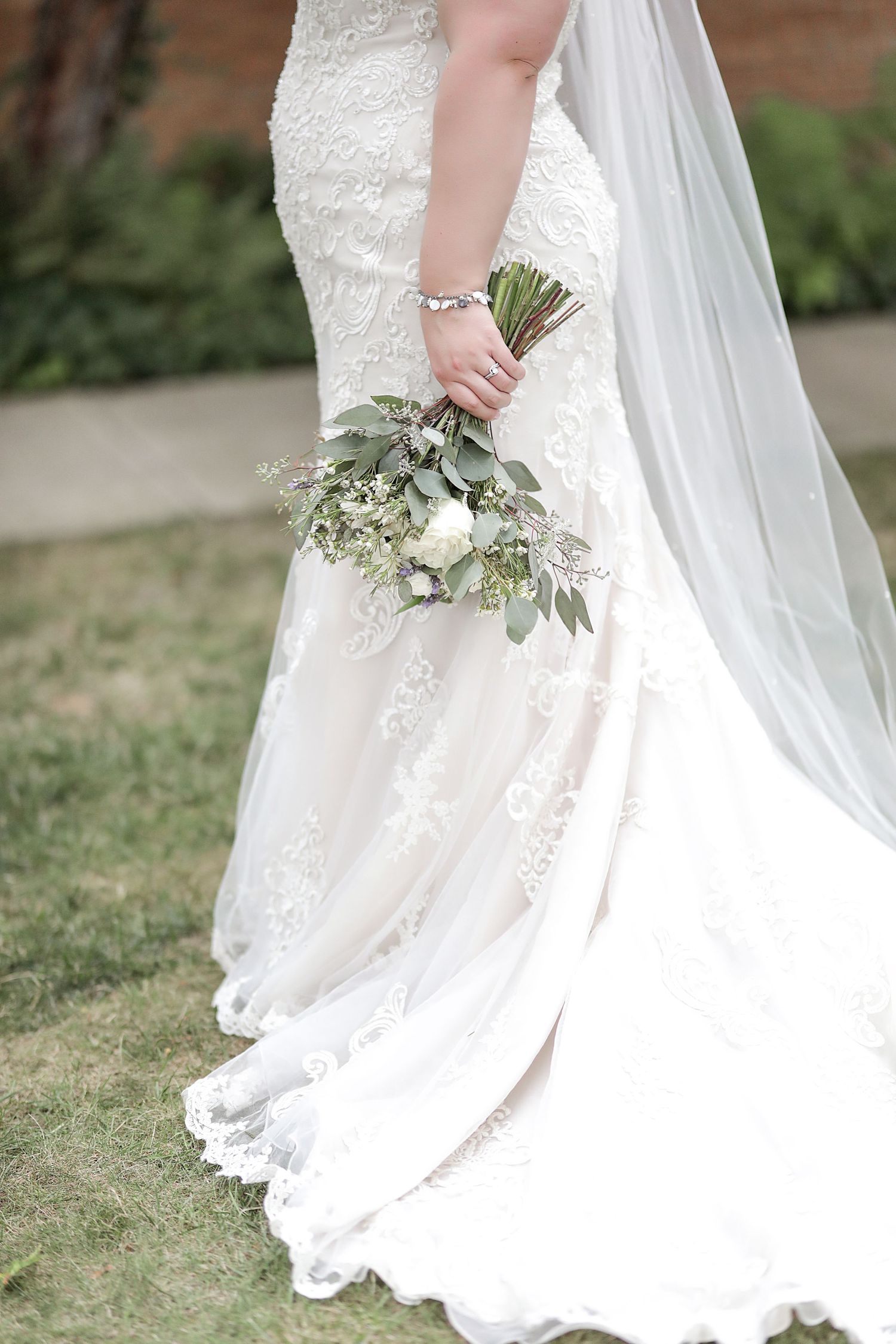 Close-up details of a white lace wedding dress train and veil flowing across green grass with delicate floral bouquet.