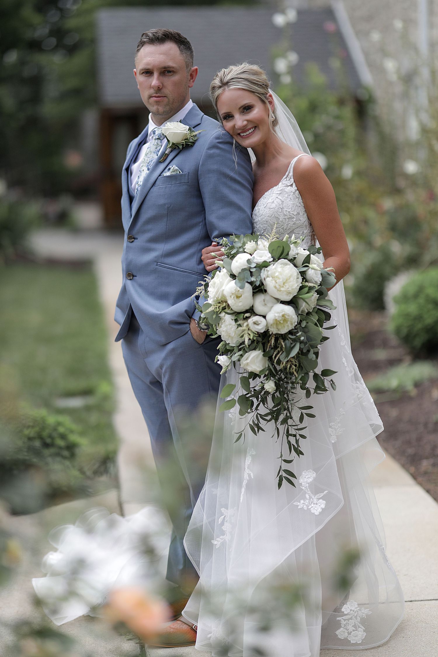 A wedding couple poses together outdoors with a stunning white floral bouquet.