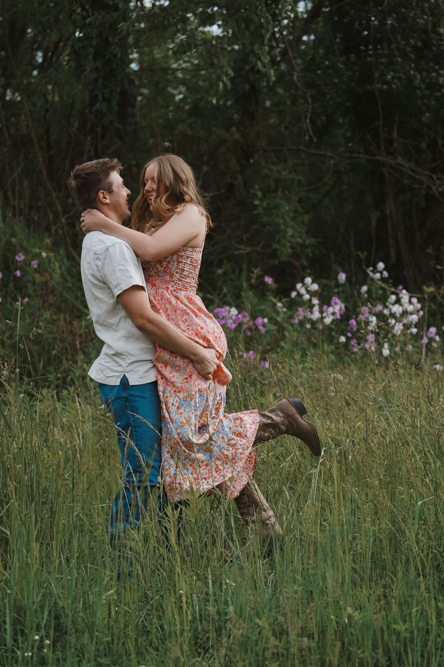 A romantic couple embraces in a wildflower meadow as golden sunlight filters through trees in the background.