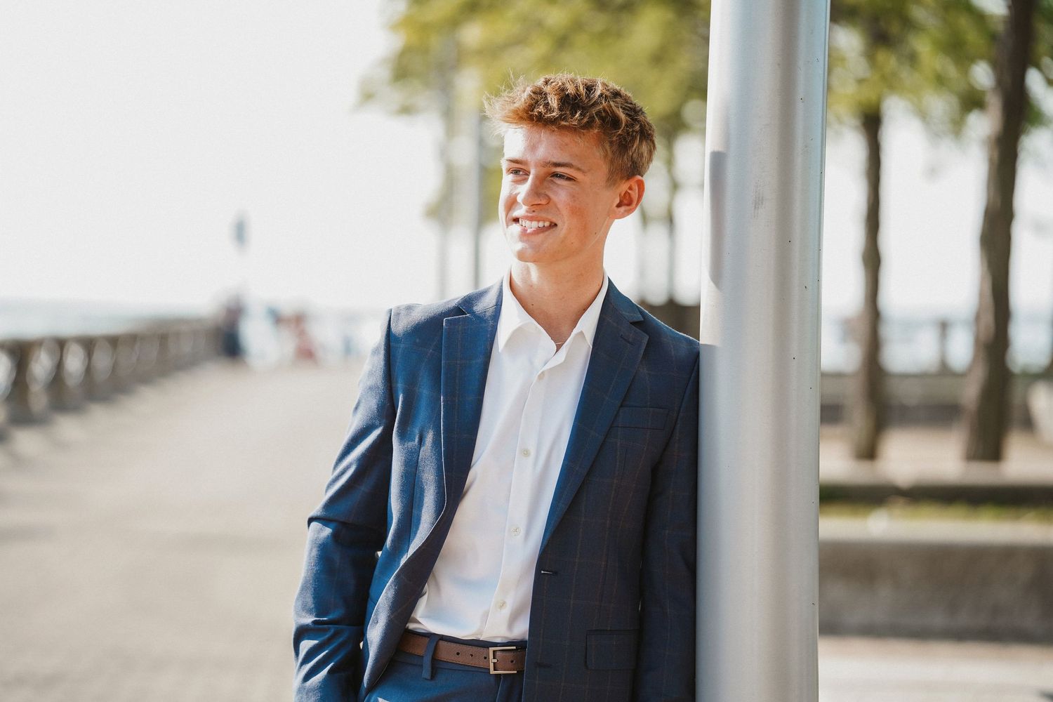Young person in a navy blue suit and white shirt smiles while standing near a waterfront walkway with palm trees.