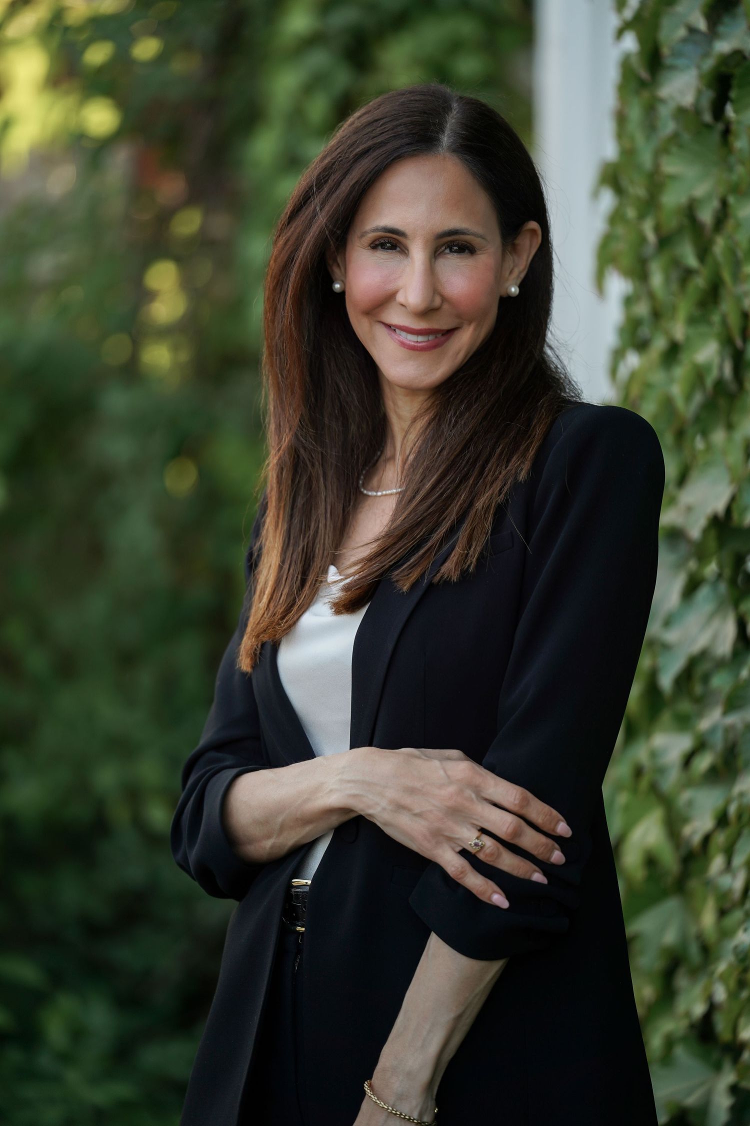 Professional portrait of someone in a black blazer and white top standing against greenery and natural lighting outdoors.