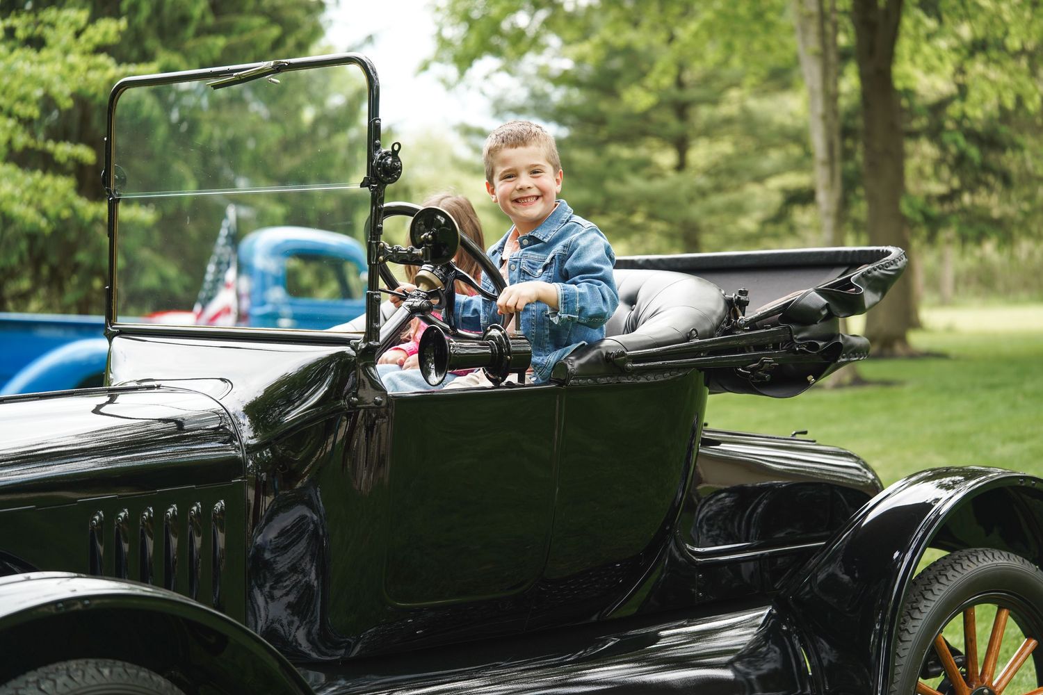 A young child rides in a restored vintage black Model T Ford automobile on a sunny day in a park setting.