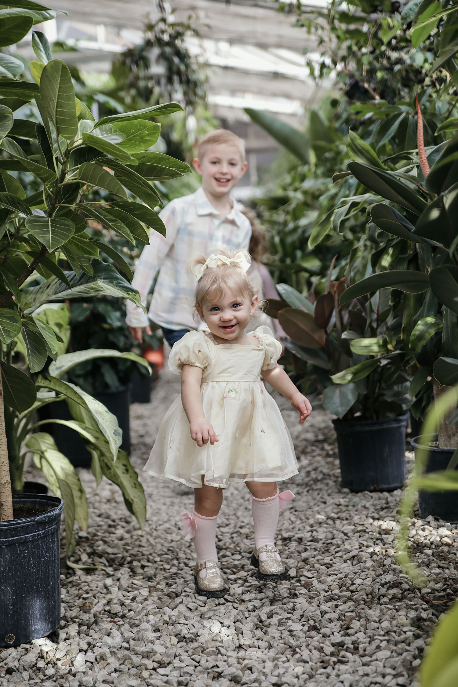 Young children in light-colored outfits play and explore among potted plants and greenery in a garden nursery setting.