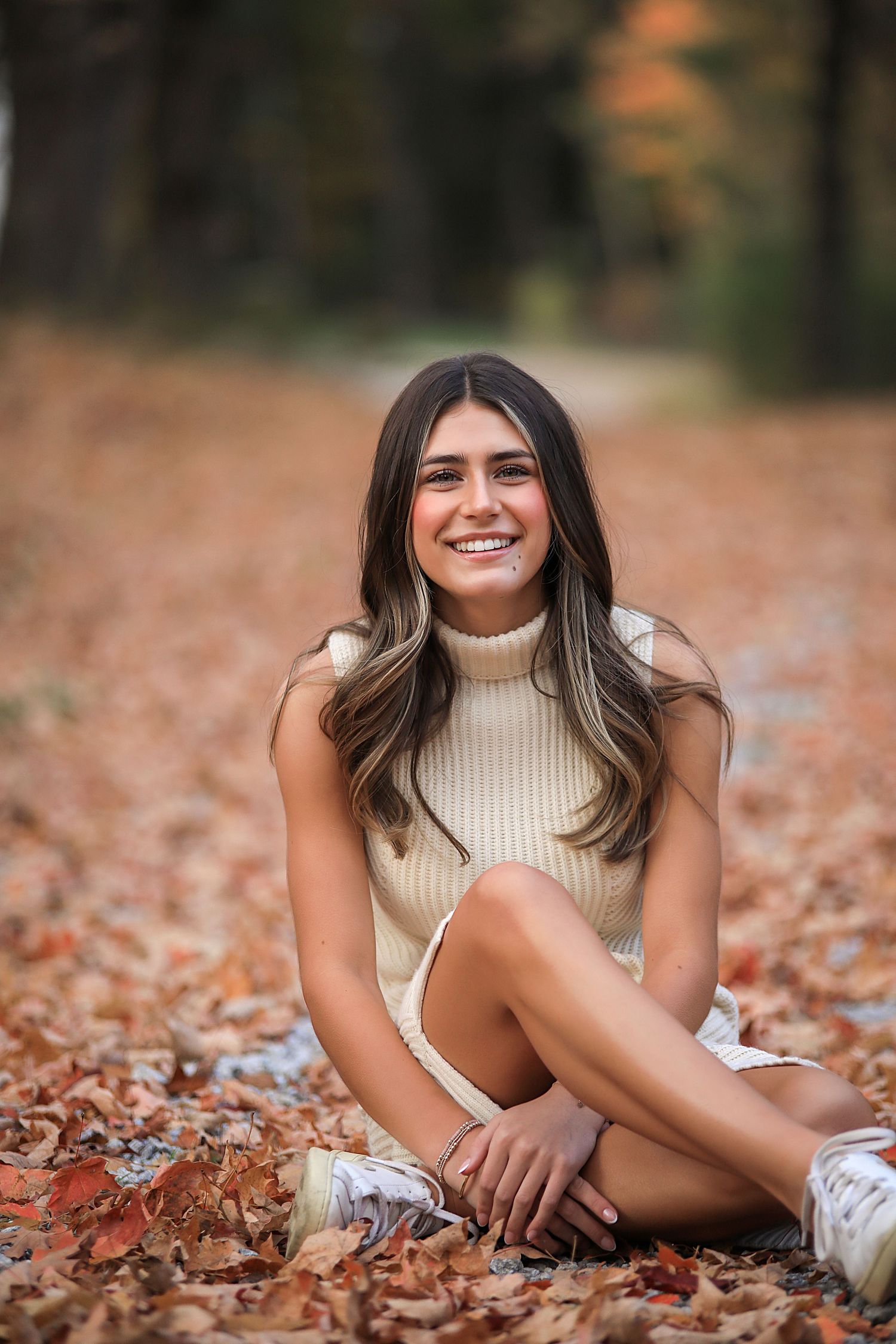 Person in cream turtleneck sweater sits on autumn leaves wearing white sneakers in outdoor portrait setting.