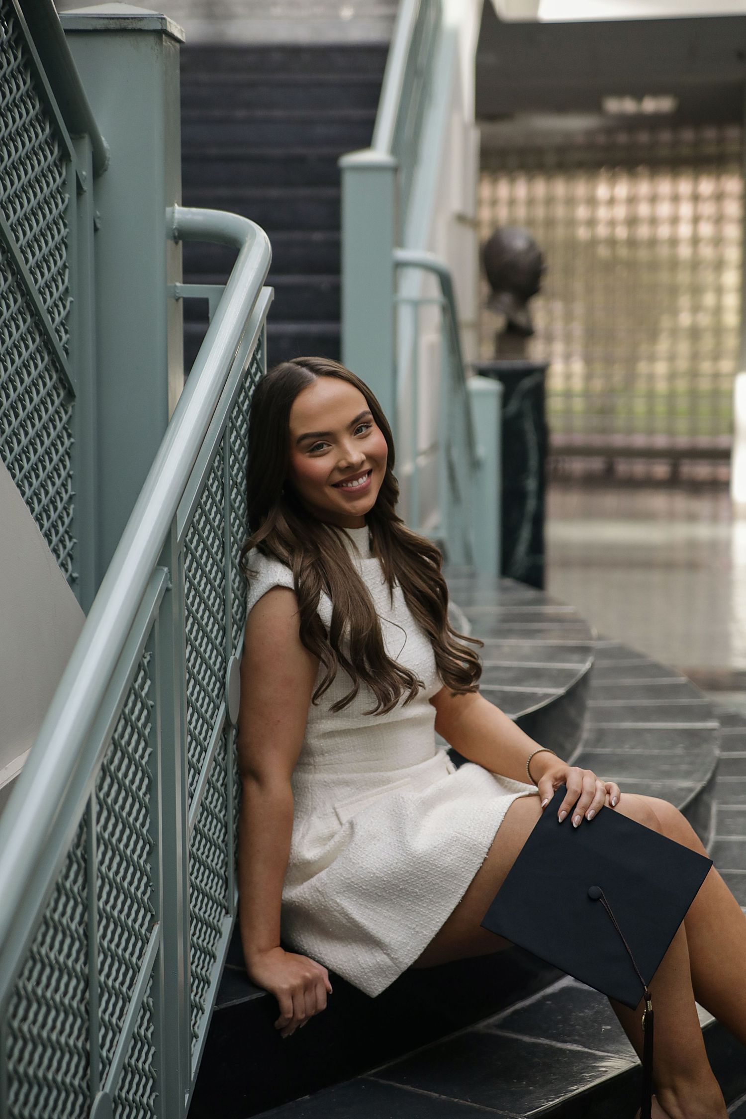 A person in a white sleeveless top and dark skirt poses against an industrial-style railing in an urban setting.