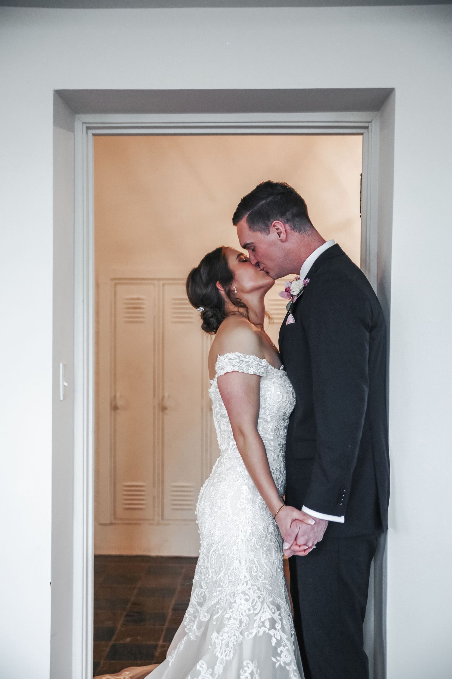 A romantic wedding couple shares an intimate kiss in a doorway, with the bride wearing a lace off-shoulder gown.
