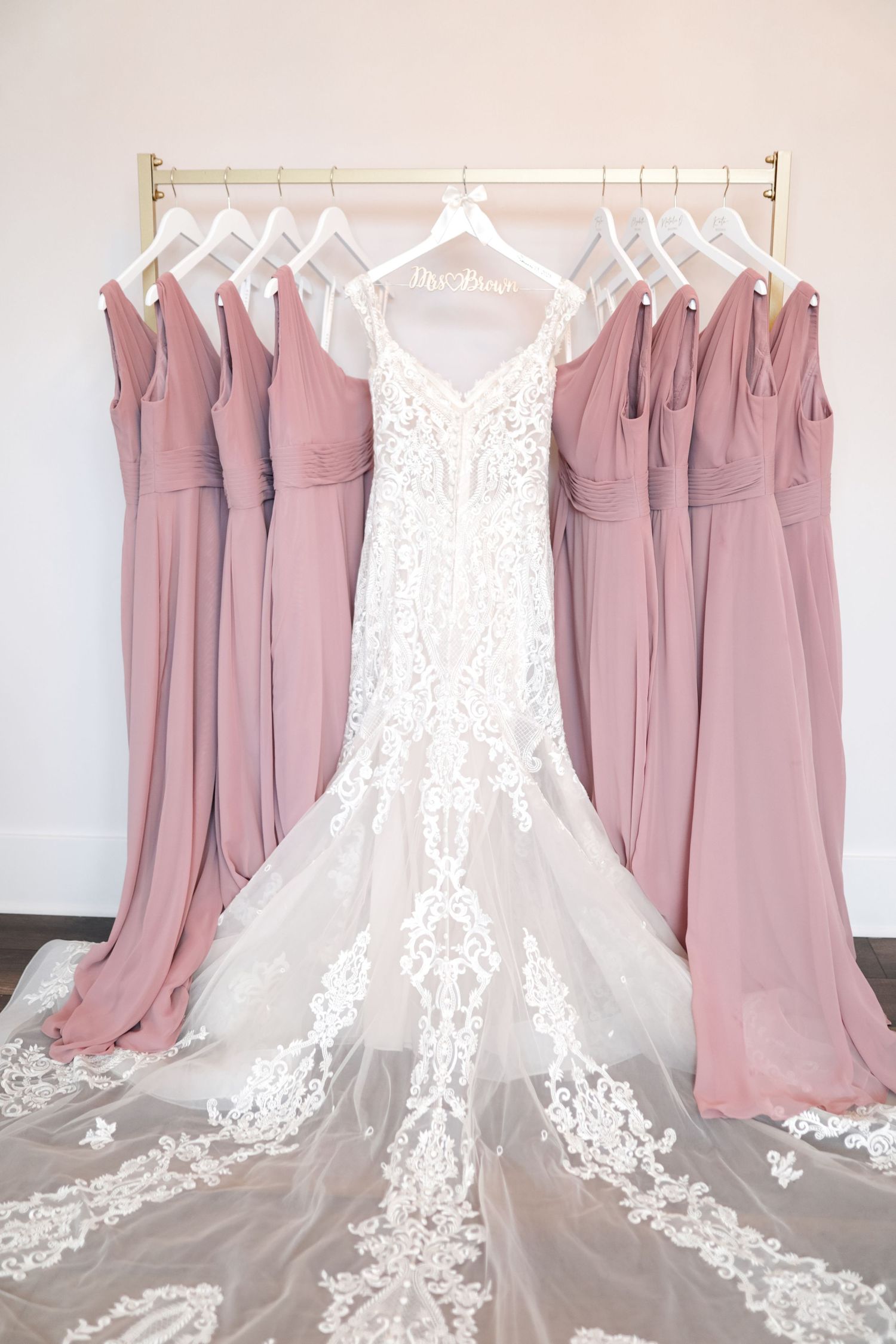 A white lace wedding gown with a cathedral train surrounded by dusty rose bridesmaid dresses on hangers.