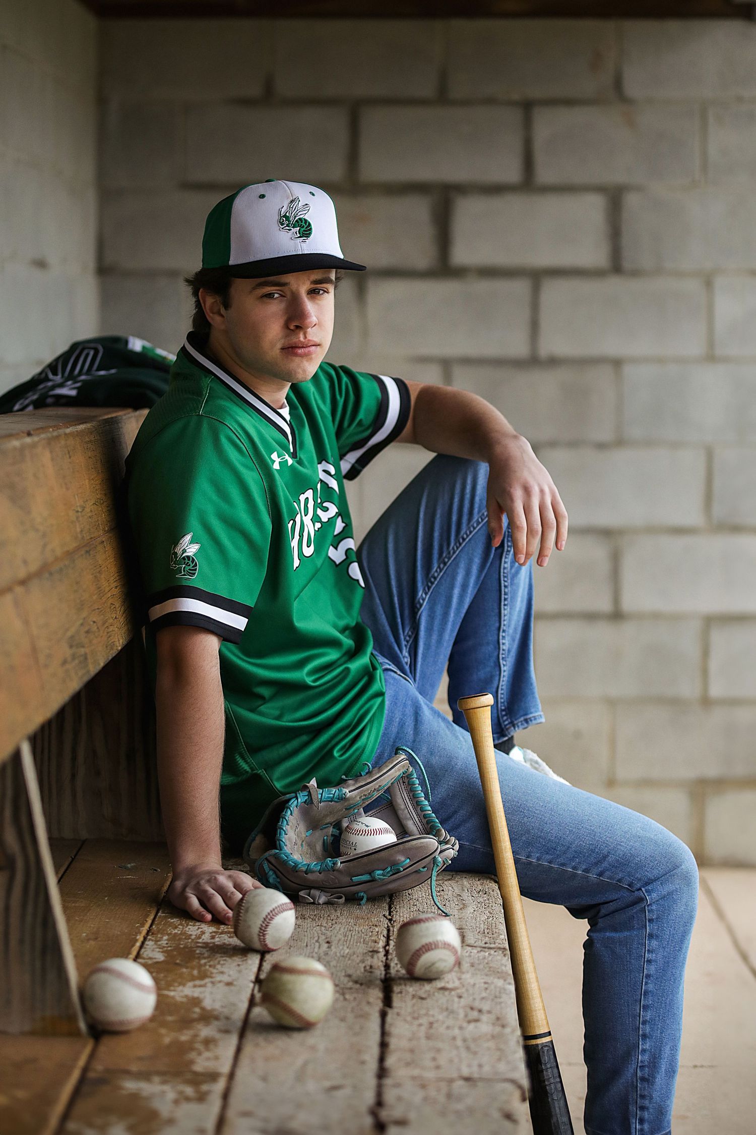 Baseball player in green jersey and jeans sits on concrete ledge with baseball bat and baseballs in casual pose.