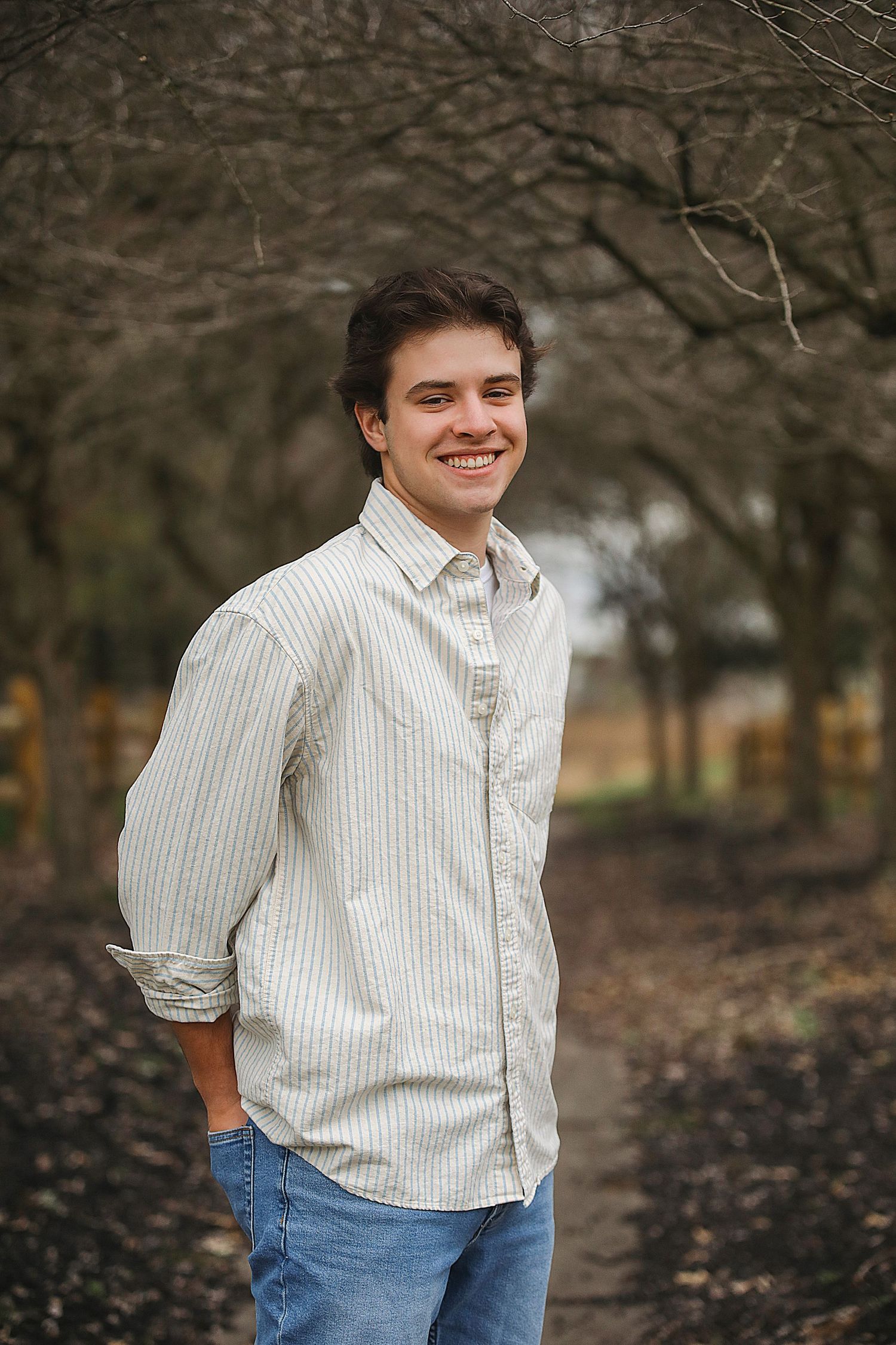 A person wearing a white pinstripe button-up shirt and blue jeans poses outdoors against a natural autumn background.