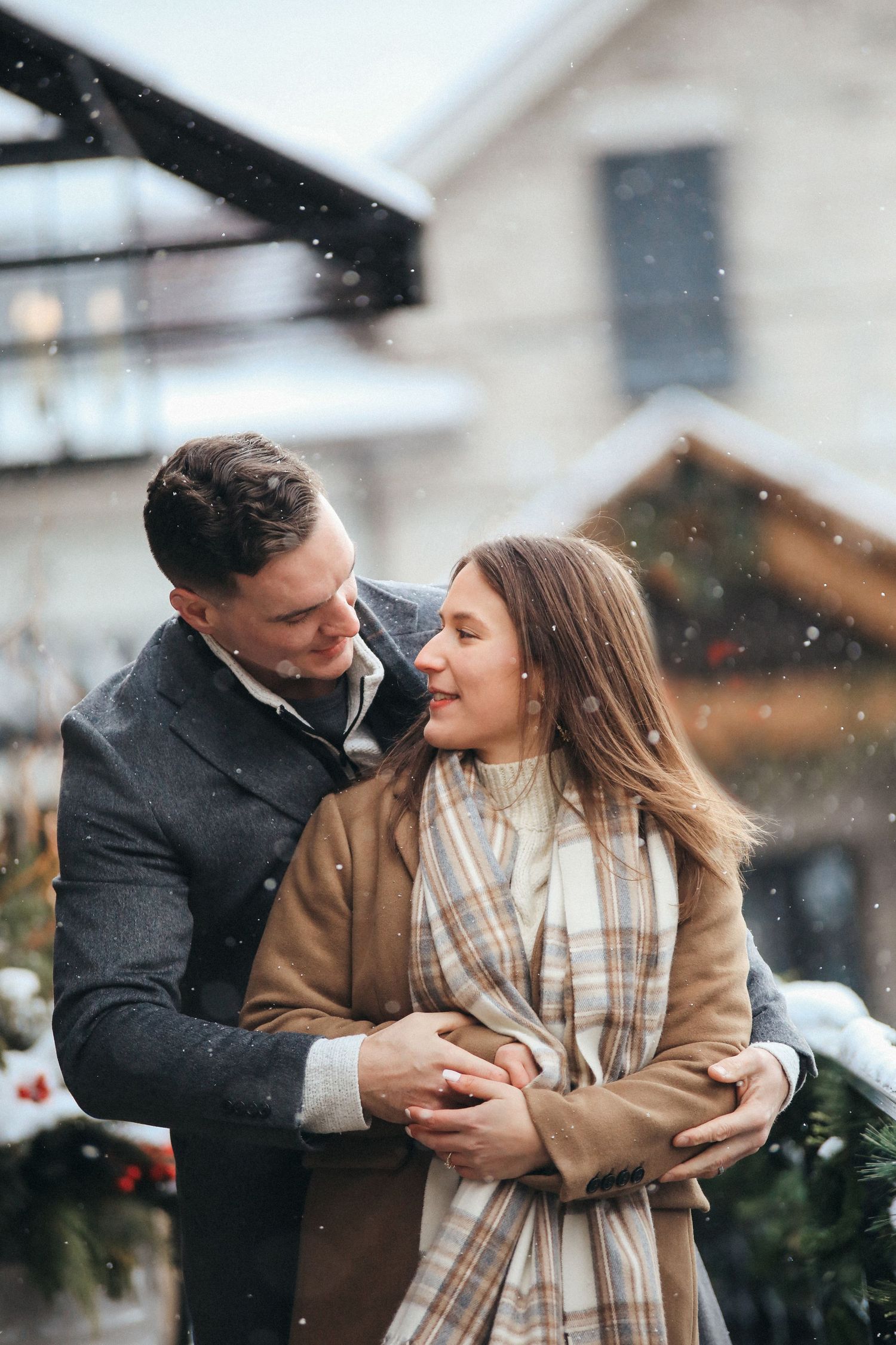 A romantic couple embraces outdoors during winter snowfall while wearing cozy coats.
