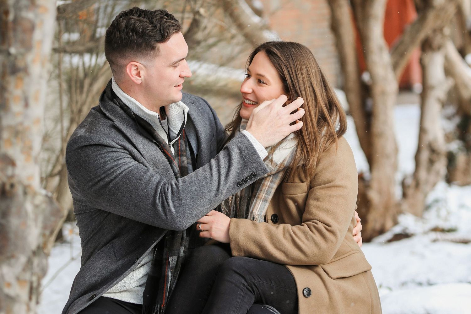 A couple shares an intimate moment while sitting together in a snowy winter setting wearing warm coats.