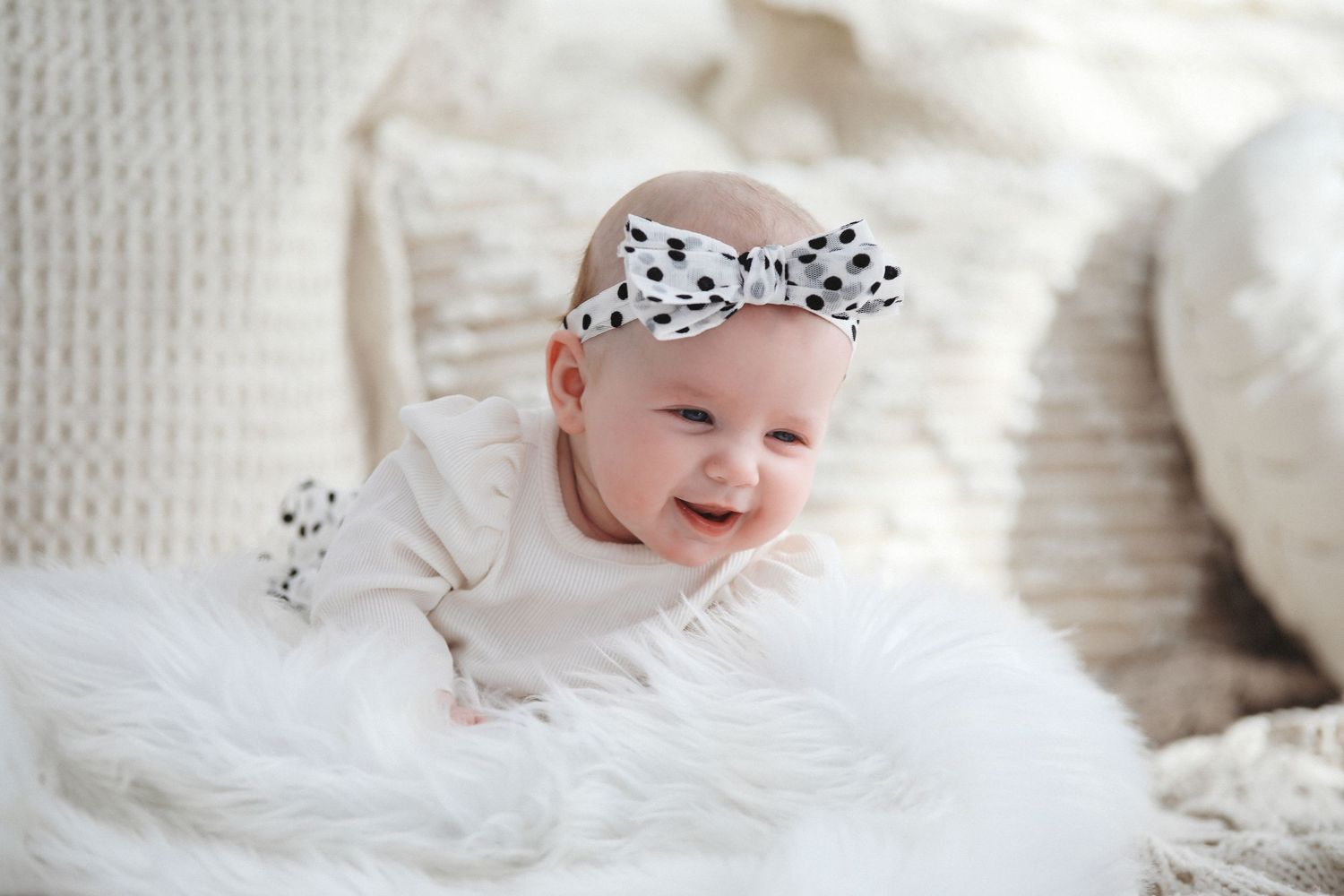 Baby wearing a polka dot bow lies on white blanket smiling with joy during newborn photoshoot.