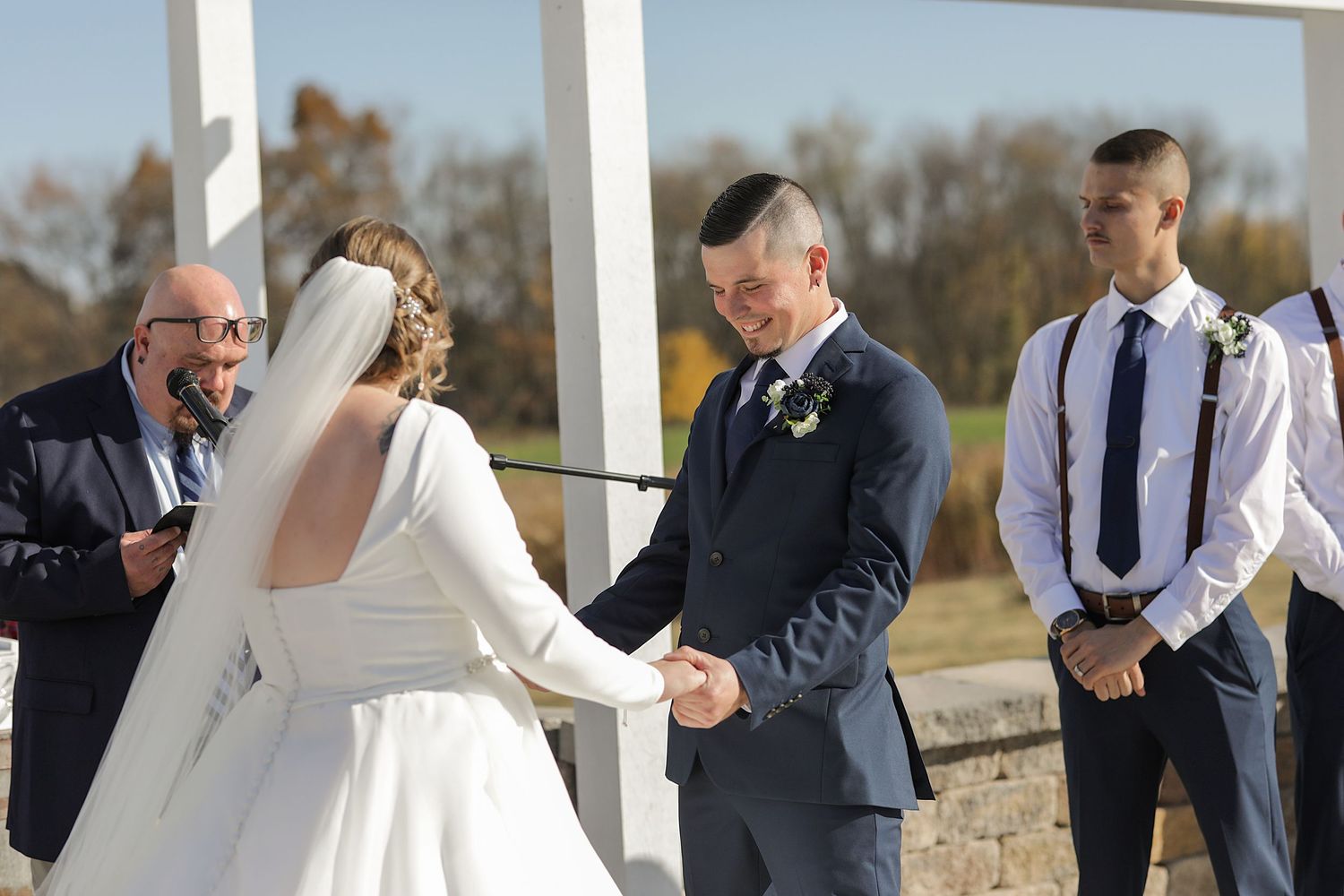 An outdoor wedding ceremony taking place under a white arch with attendants looking on.