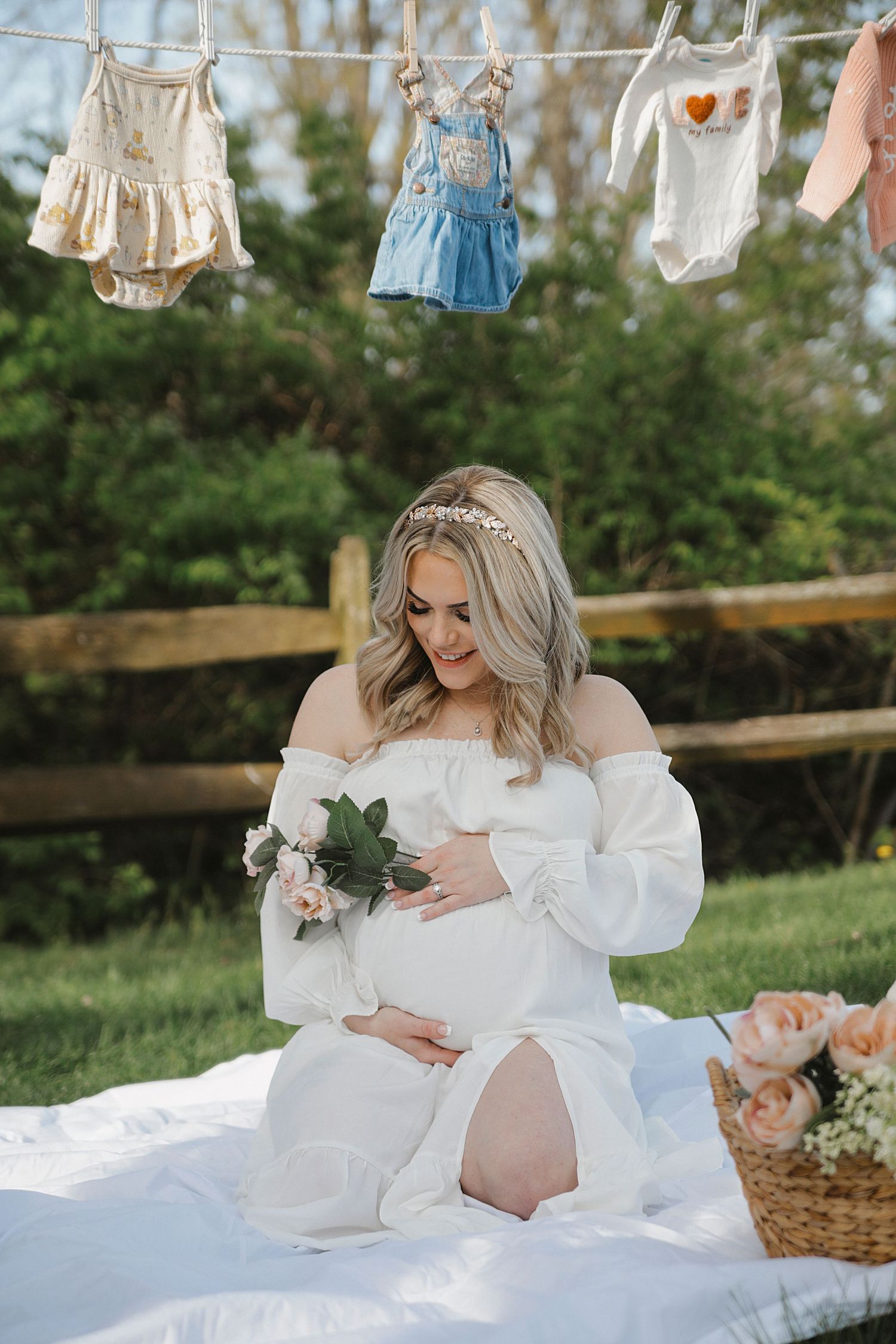 Maternity photoshoot scene with woman in white flowing dress reclining on blanket outdoors with hanging clothes in background.