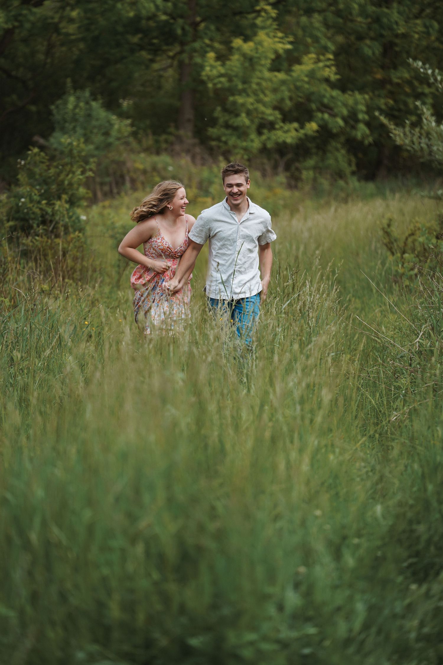 A couple runs joyfully through a tall grass meadow surrounded by trees on a summer day.