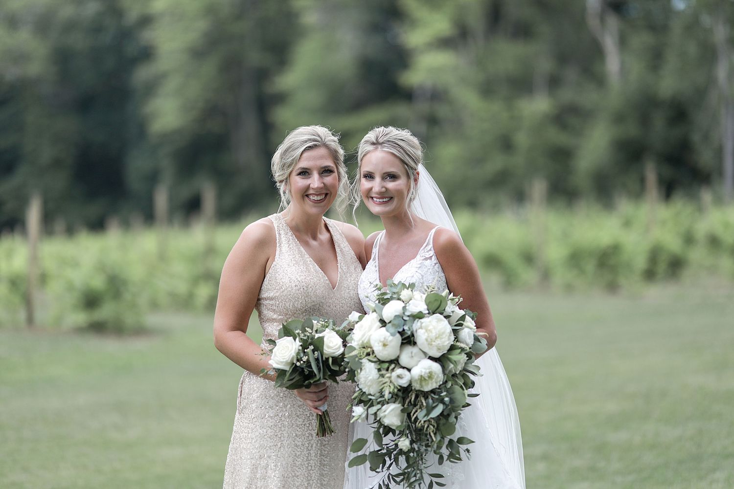 Two wedding party members pose with white rose bouquets against a lush green backdrop.