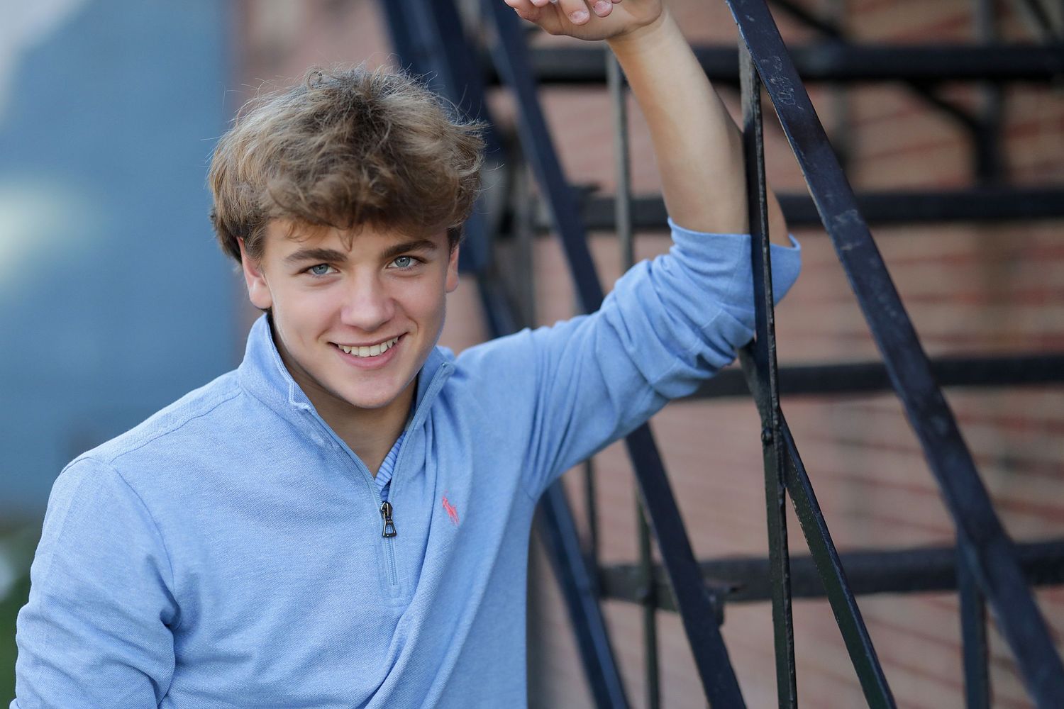 Student in light blue quarter-zip pullover smiles while leaning on outdoor metal railing on steps.