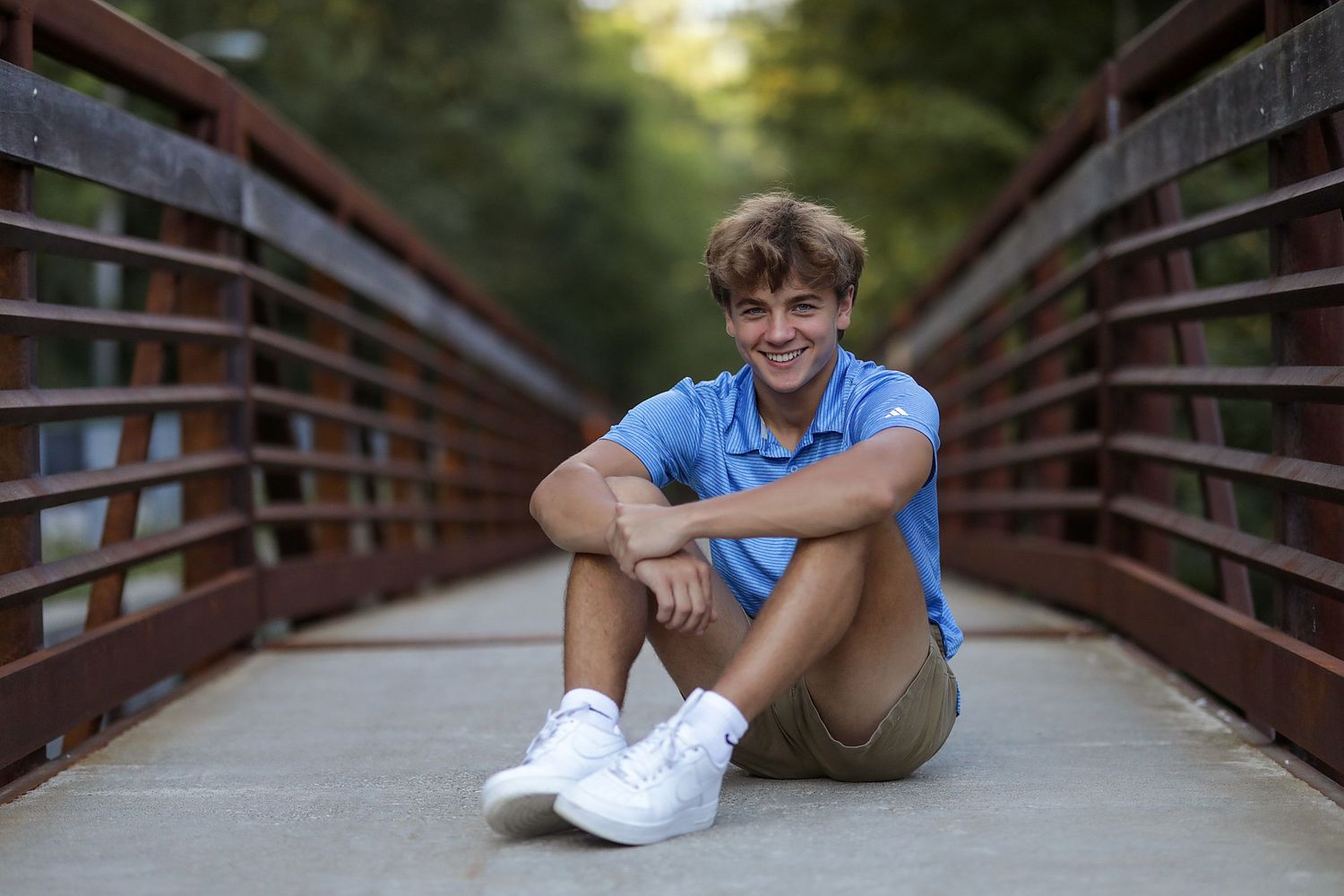 A young person in a blue shirt and khaki shorts sits casually on a wooden bridge on a sunny day.