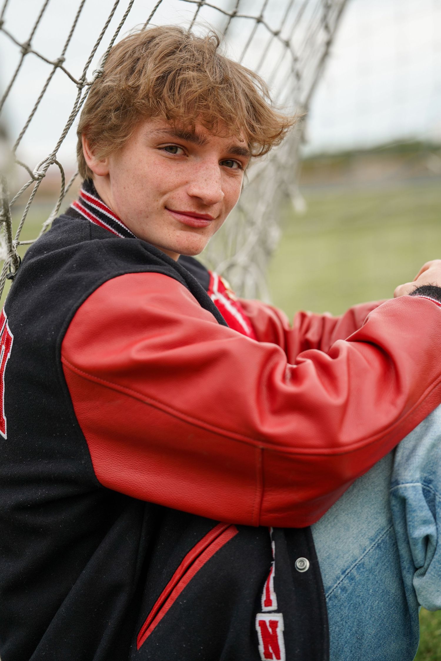 A person in a red and black varsity jacket leans against a chain-link fence on a cloudy day.