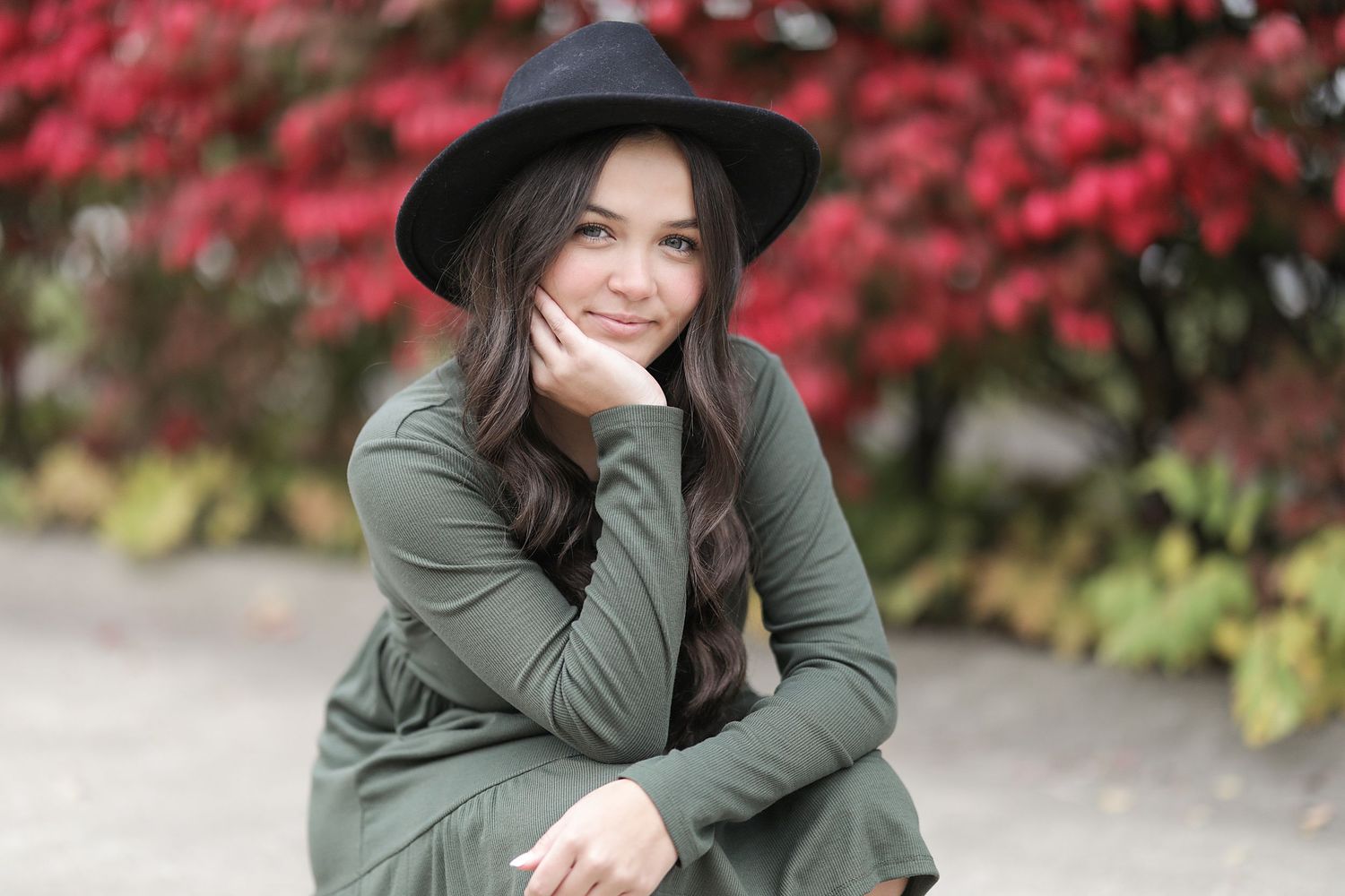 Casual portrait outdoors with dark hat and olive sweater against vibrant red autumn foliage background.