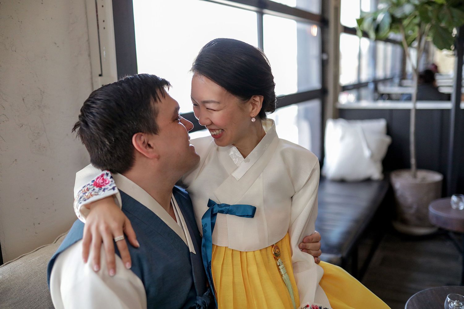 A joyful multicultural couple embraces and laughs together, with one wearing traditional hanbok dress.