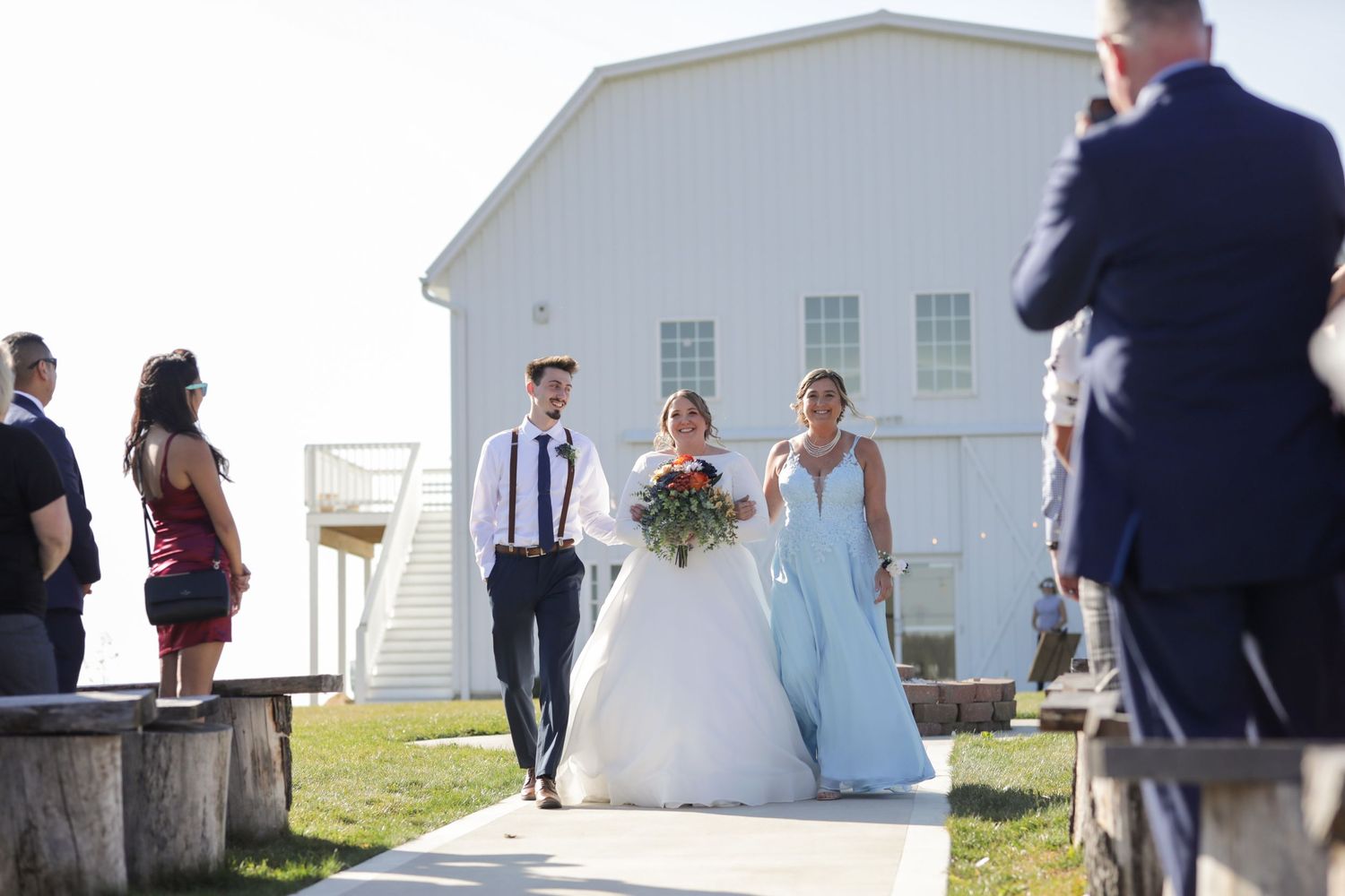 A wedding party walks along a sun-lit path outside a white barn venue on a bright summer day.