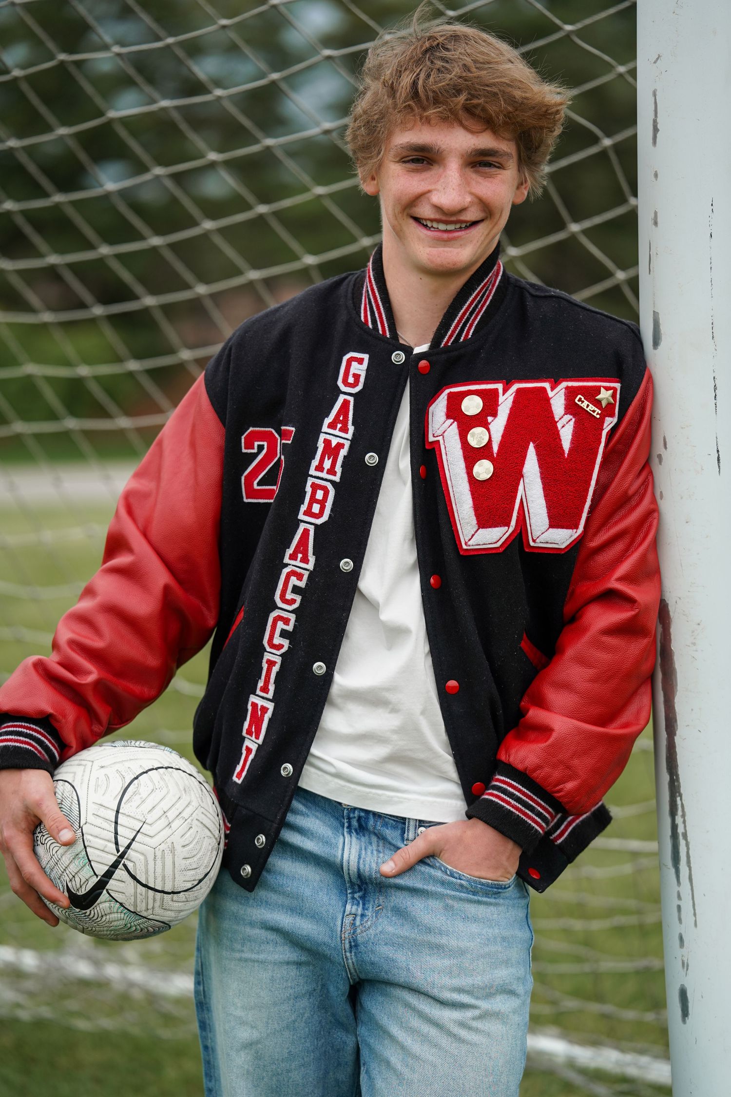 High school athlete in red and black letterman jacket holds volleyball by soccer goal wearing jeans and casual attire.