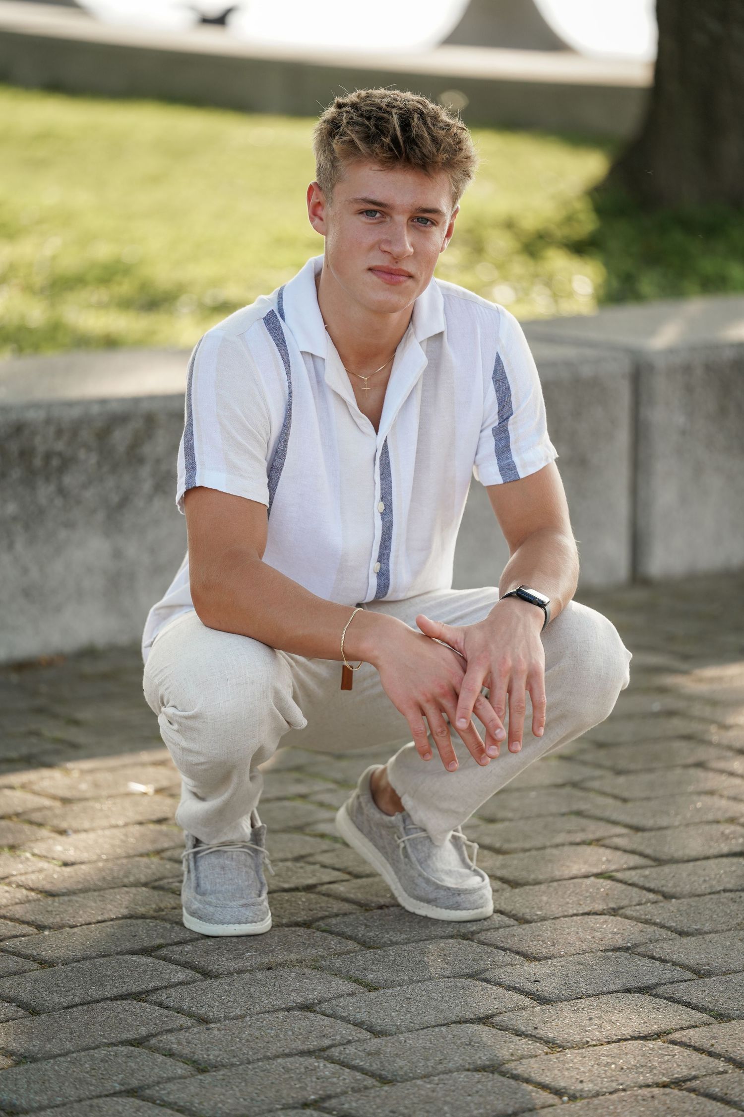 Young person in white button-down shirt and cream pants crouches on brick pavement in natural lighting.