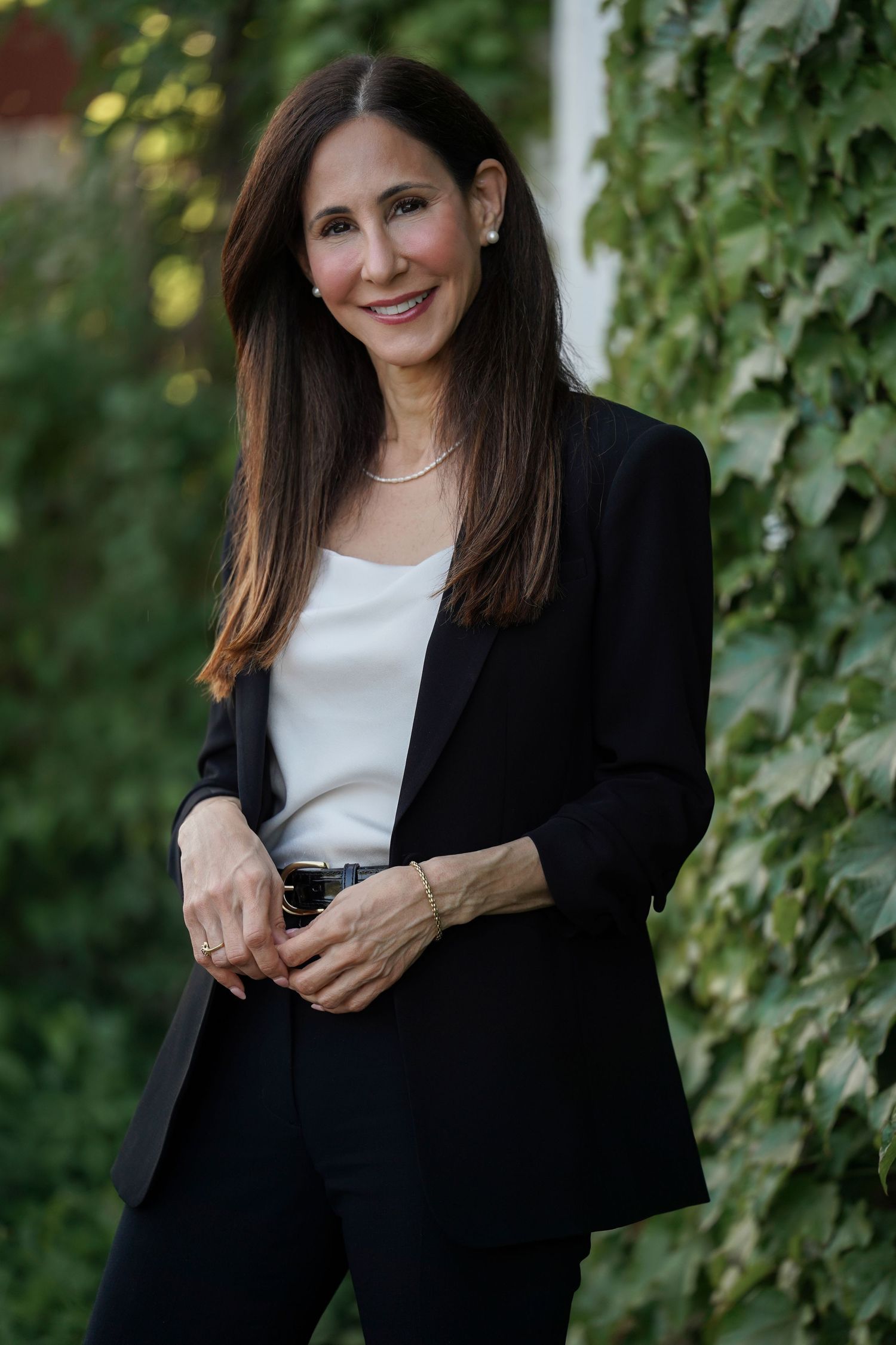 Professional portrait of person in black blazer and white top standing against ivy covered wall outdoors.