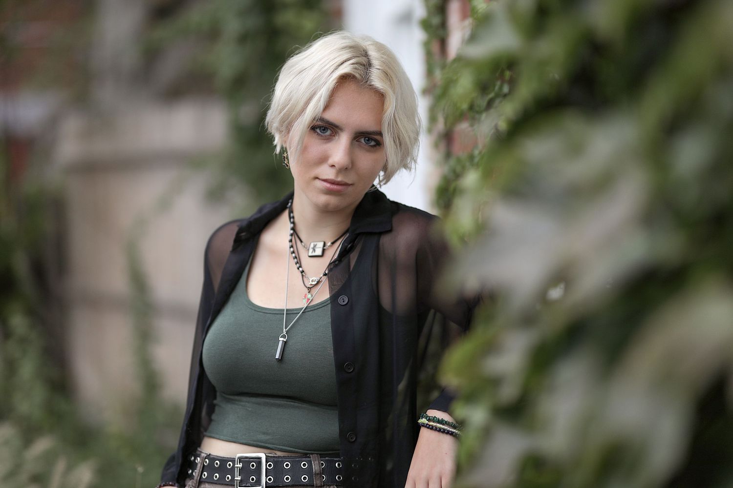 A person with platinum blonde hair poses outdoors wearing a sheer black cardigan over a dark top.