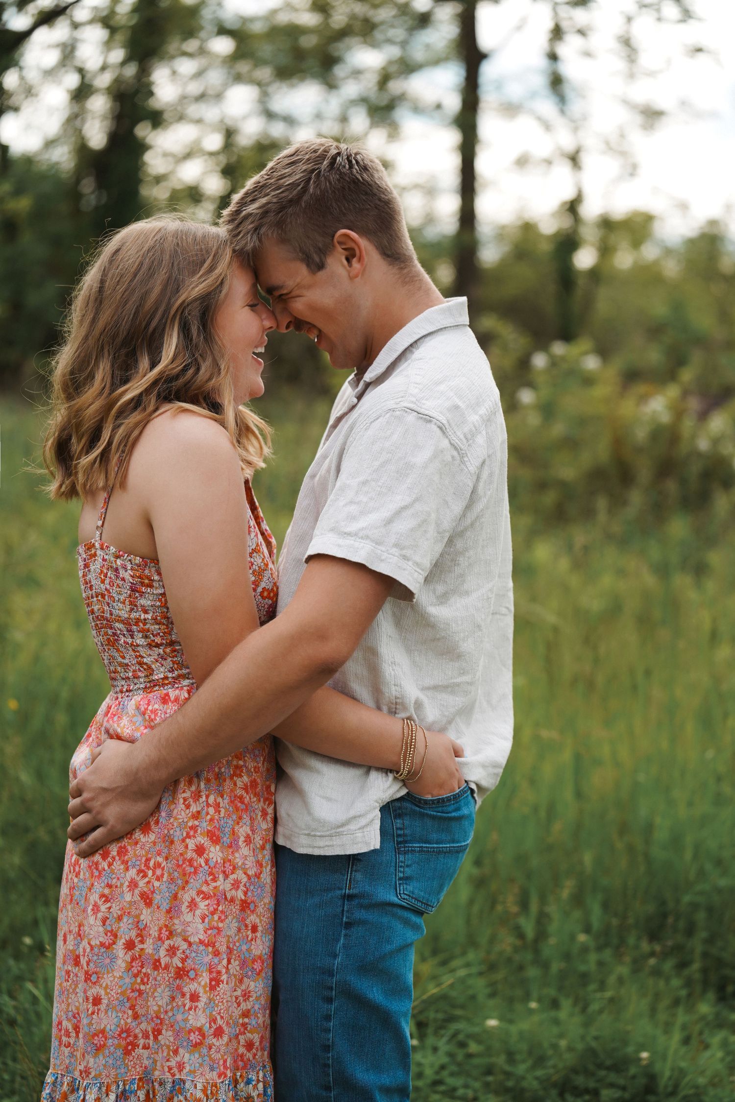 A romantic couple in casual attire embraces tenderly in the golden light of a meadow at sunset.