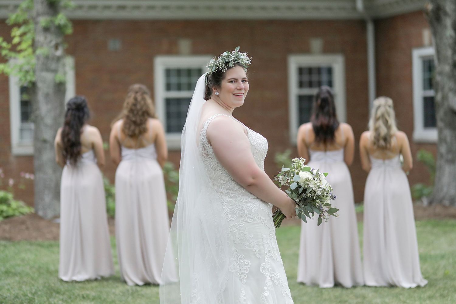 Bride in a lace wedding dress with flower crown stands in front of bridesmaids wearing blush gowns on a grassy lawn.