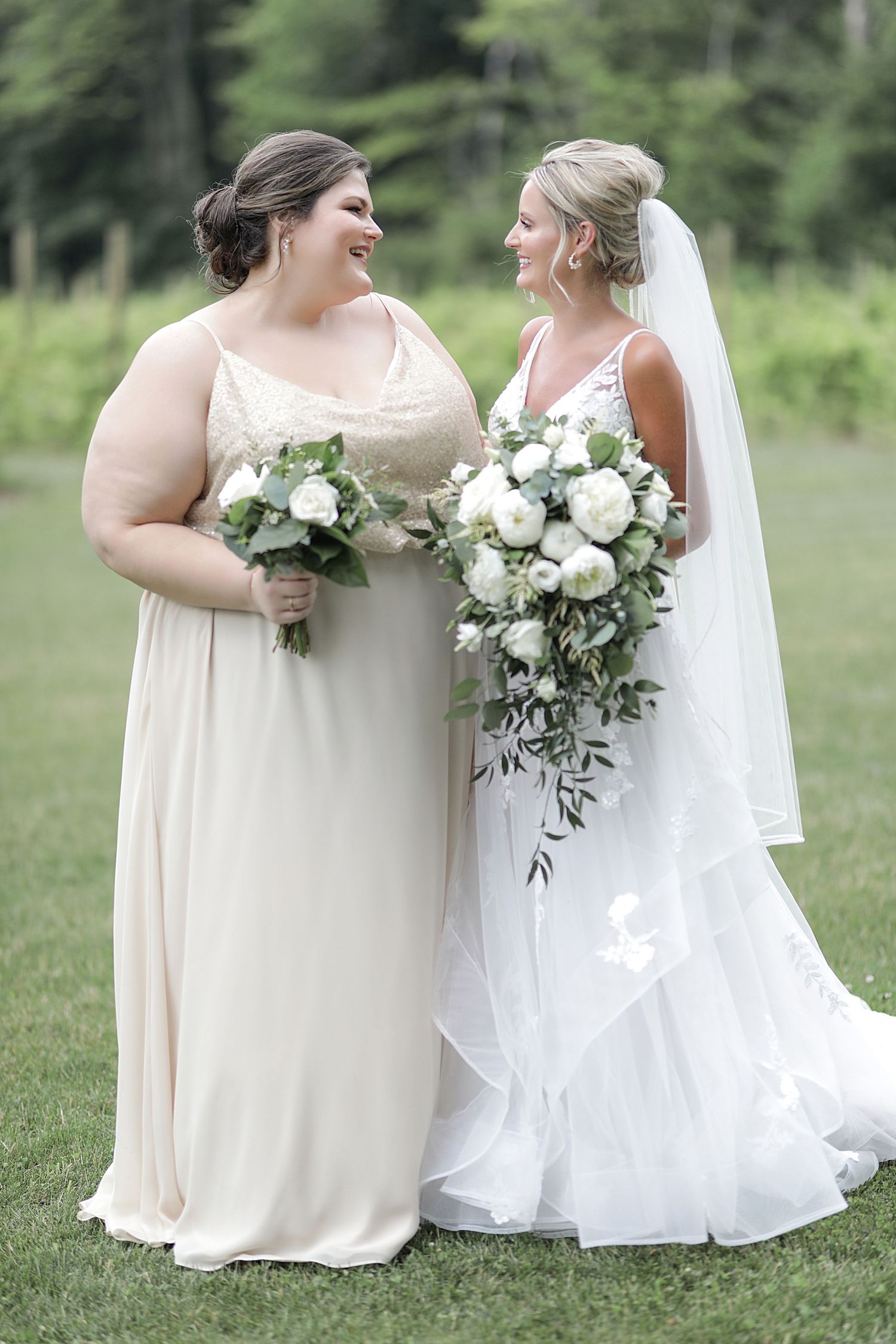 Bride and bridesmaid pose with white rose bouquets on grassy lawn.