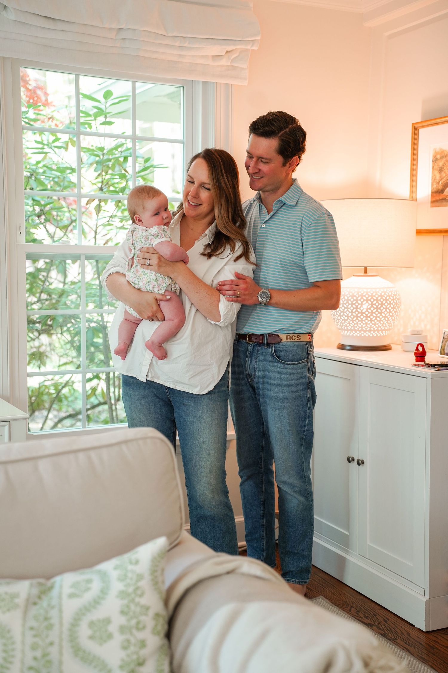 A couple stands by a bright window in a cozy living room while holding a baby, creating a warm family moment at home.